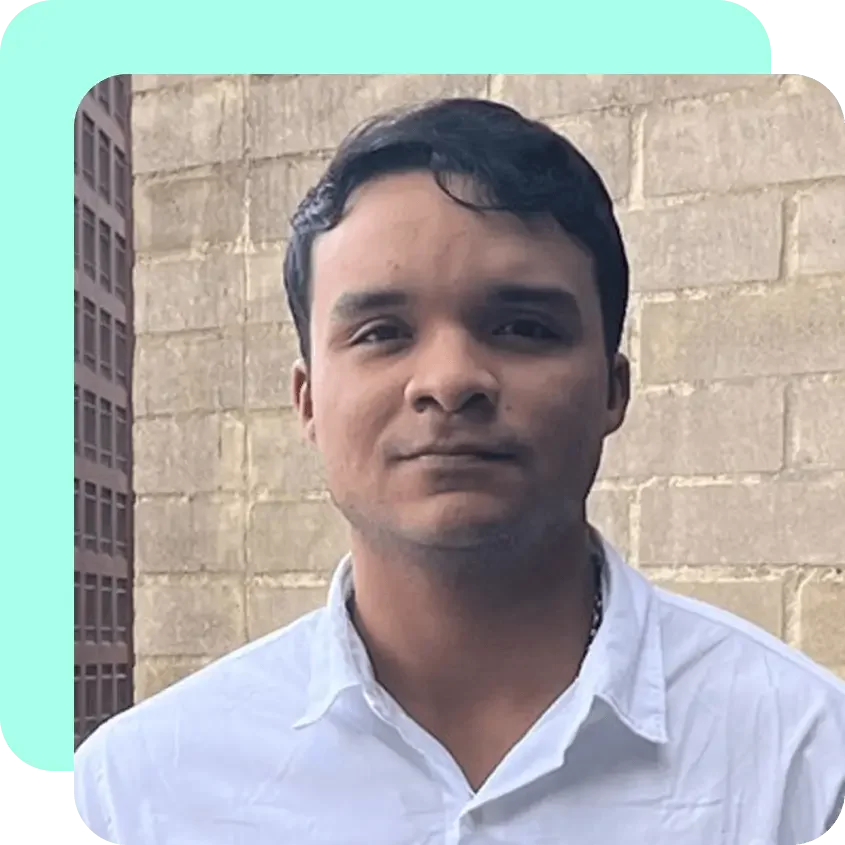 A young man with short dark hair, wearing a white button-up shirt, standing outdoors in front of a brick wall with an urban building in the background.