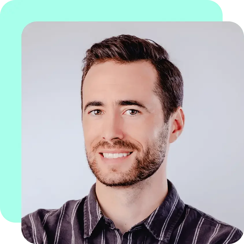 Close-up portrait of smiling man with dark hair and beard, wearing a dark plaid shirt, against a light background.