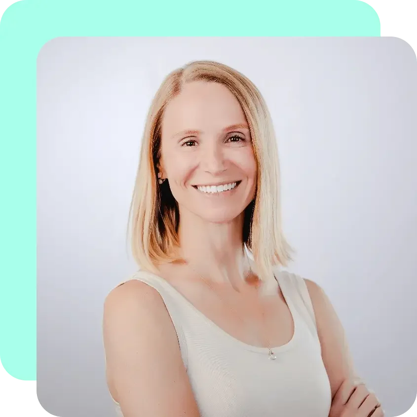 A smiling woman with blonde hair wearing a white sleeveless top and a delicate necklace, standing against a plain white background.