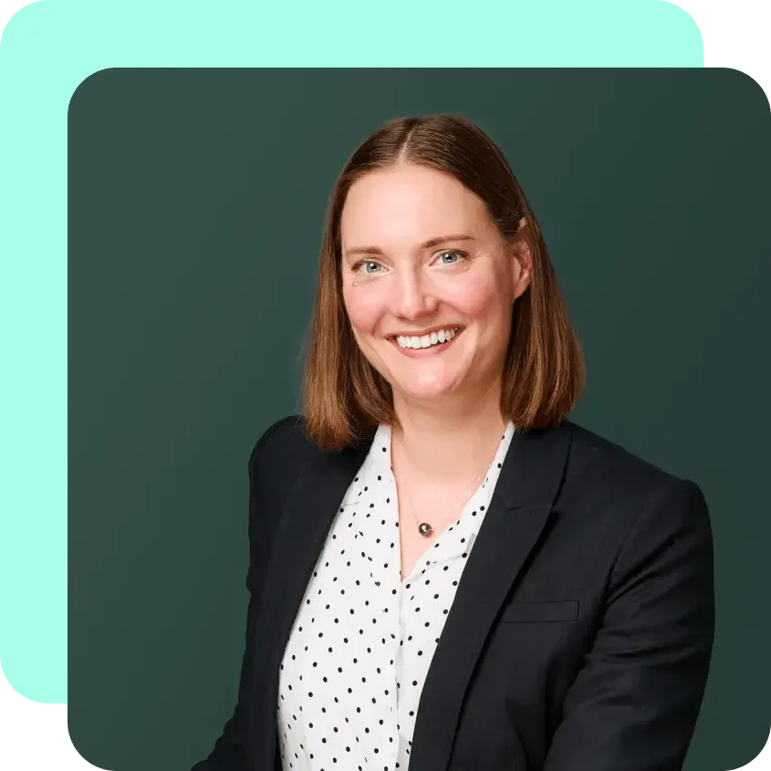 Portrait of a woman with shoulder-length brown hair, wearing a black blazer and a white blouse with black polka dots, smiling against a green background.