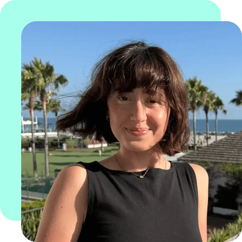 A young woman with short brown hair and bangs smiling outdoors near the ocean, surrounded by palm trees and clear blue sky.