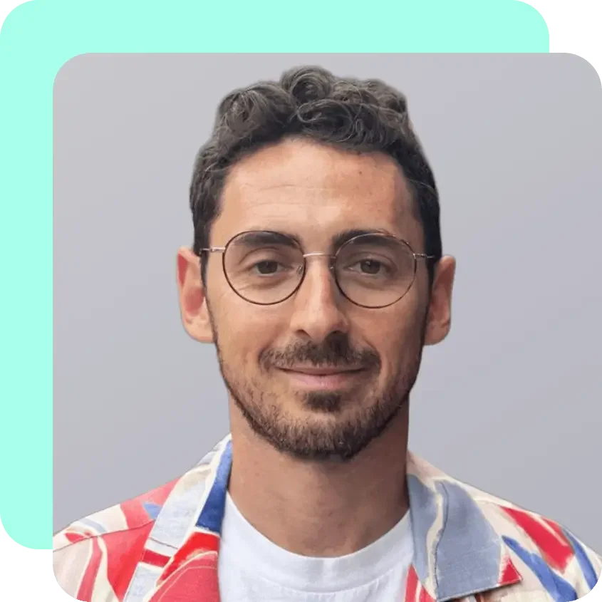 Portrait of a man with short curly brown hair, glasses, a slight beard, wearing a colorful patterned shirt, smiling against a neutral background.