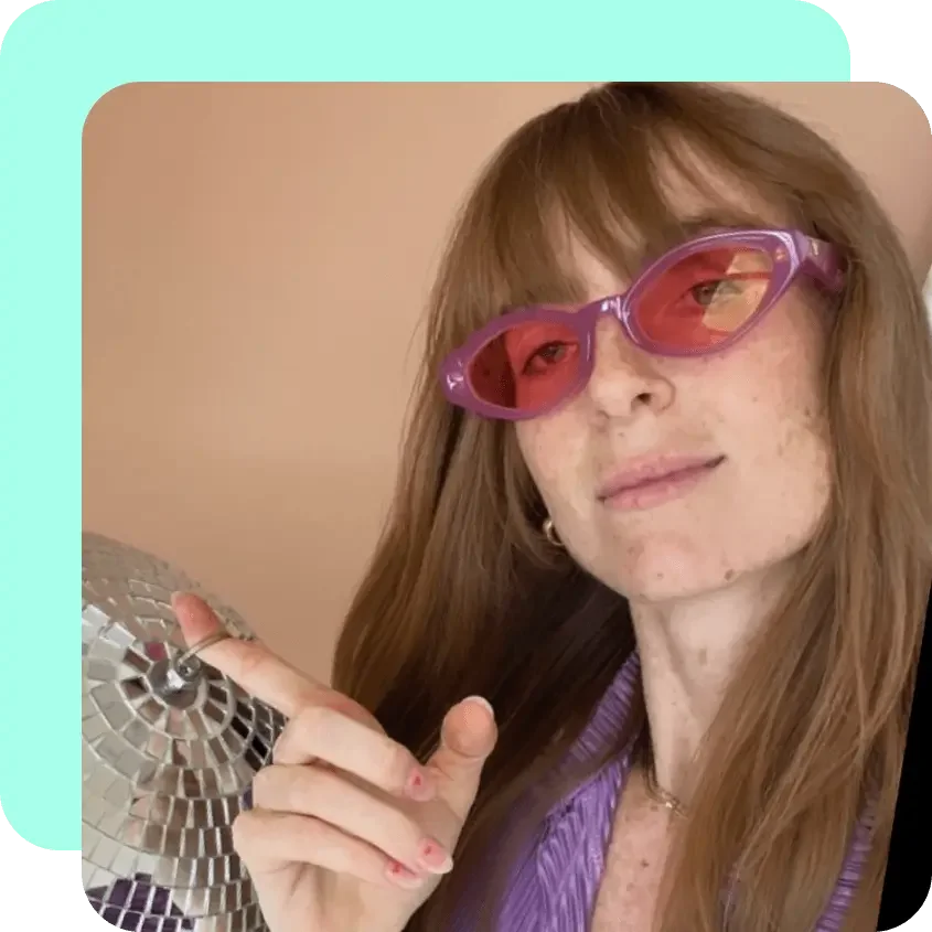 A woman with red hair and freckles wearing pink sunglasses and a purple shirt, holding a small disco ball, pointing at it with her finger.