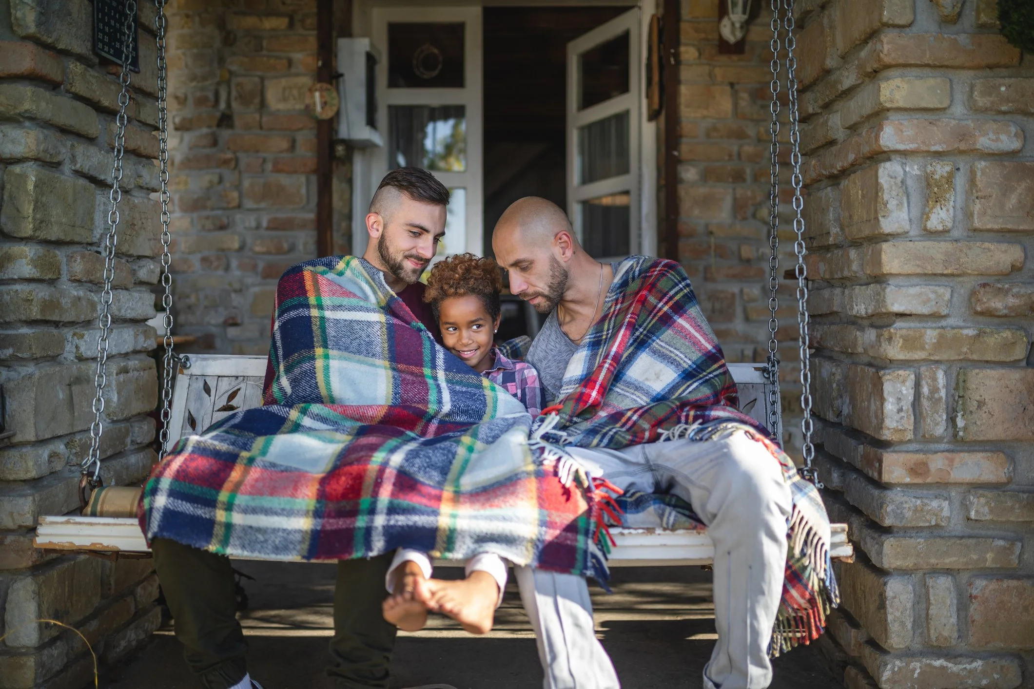 Two dads and their daughter wrapped in a blanket on a porch swing