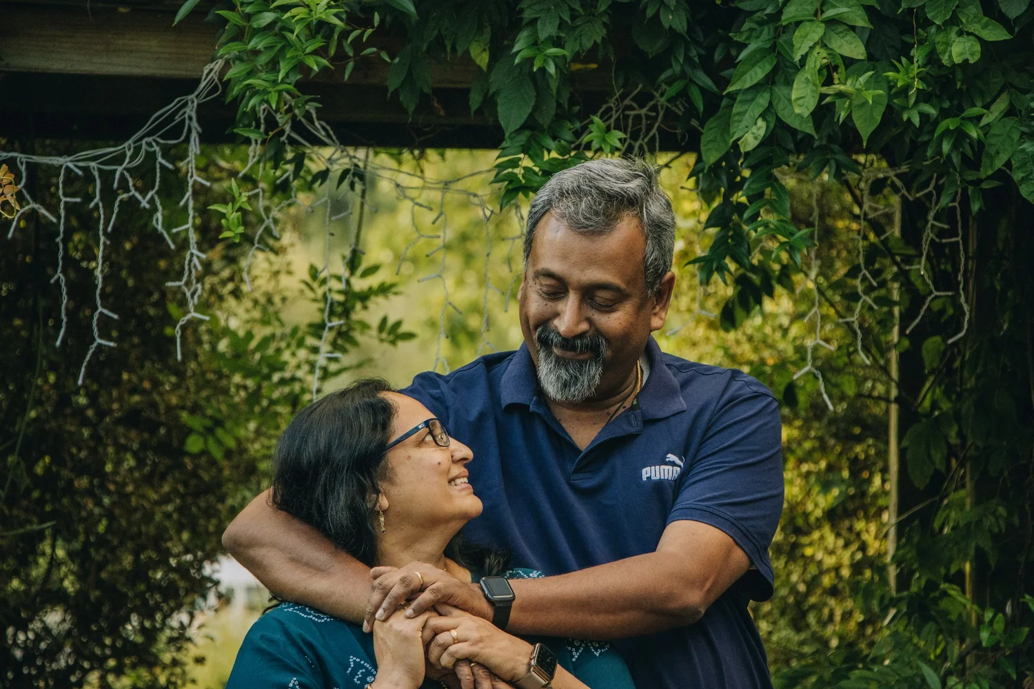 A couple in their fifties laughing together under a vine-covered trellis, his arms around her from behind