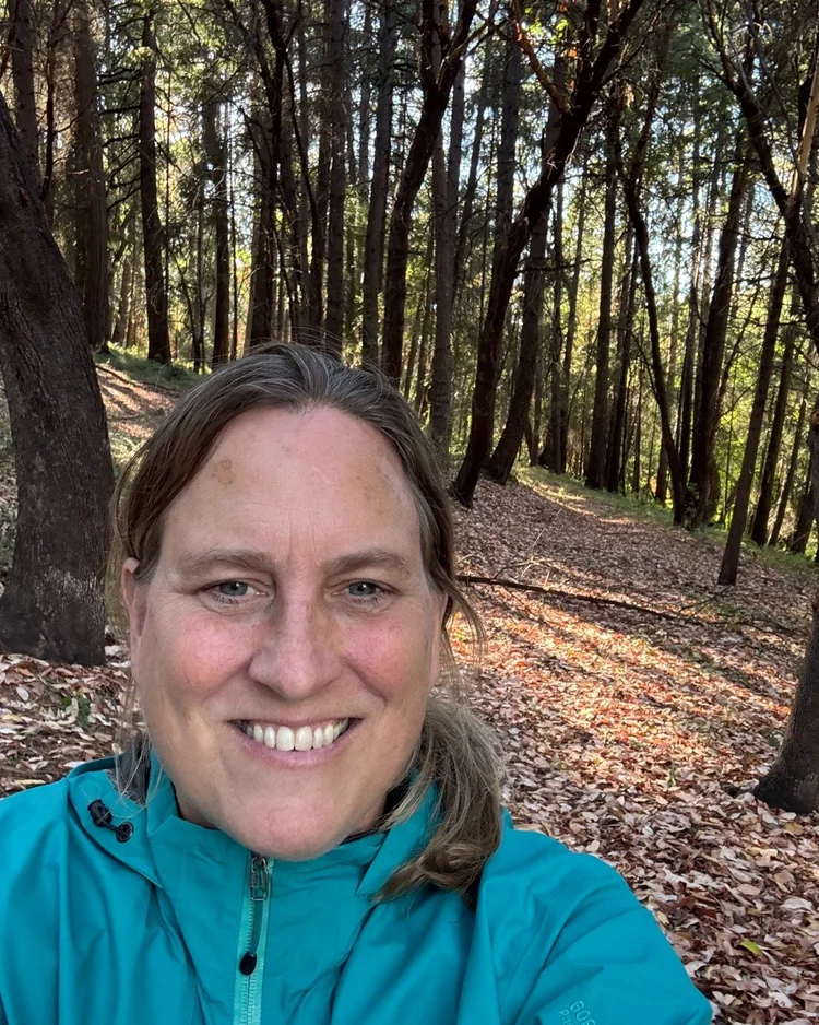 Janette Robinson smiling on a leaf-covered trail curving away through the pines