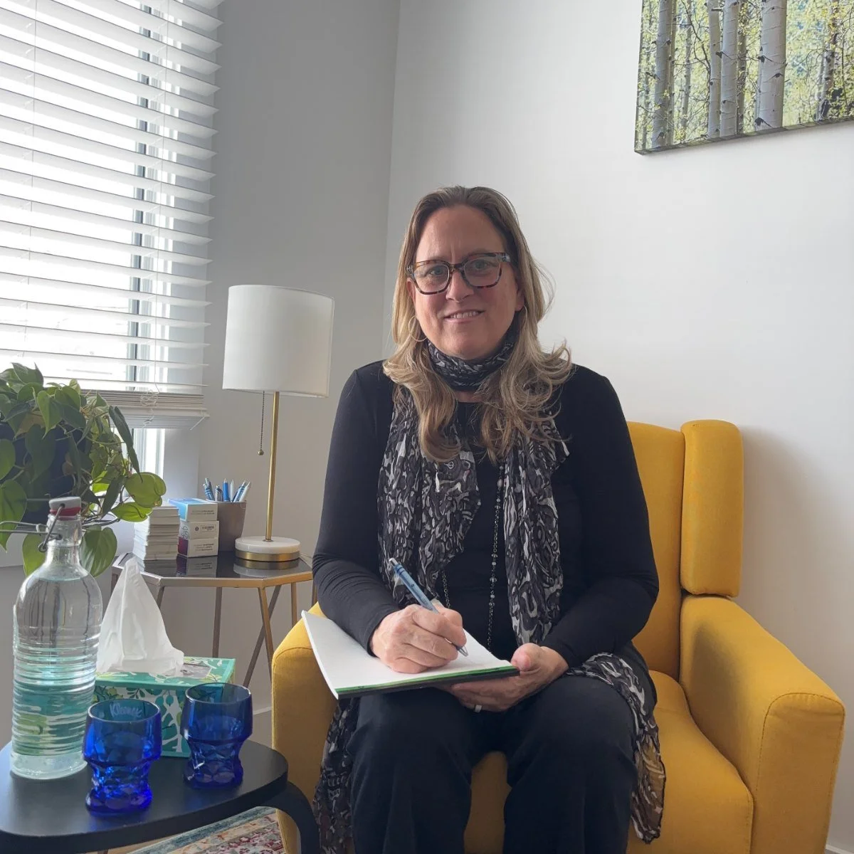 Janette Robinson seated in the yellow chair in her consulting room, notebook open, a bottle of water and two glasses on the table beside her