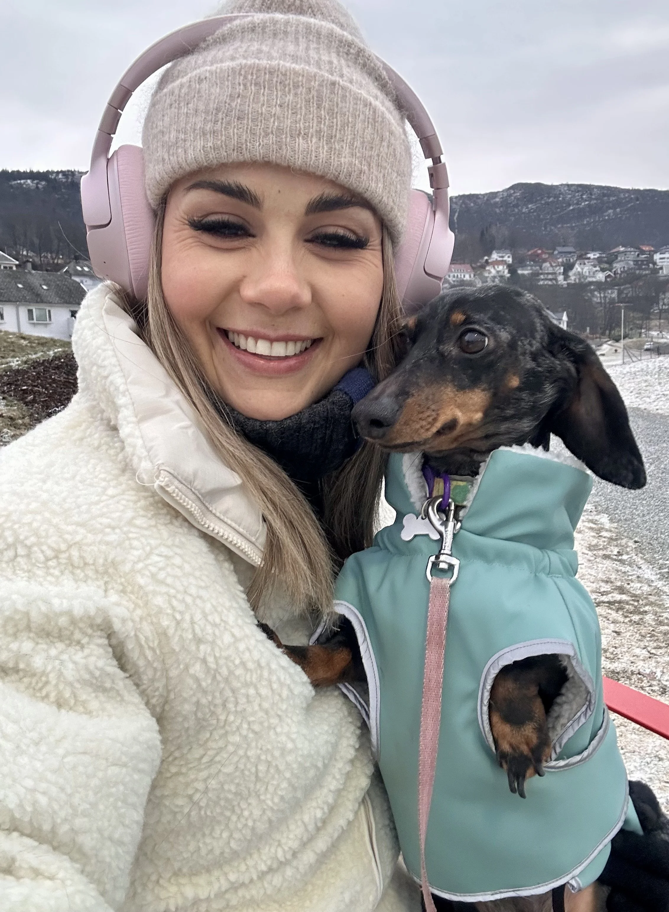 A woman smiling for a selfie with her dog in a snowy outdoor setting, wearing a beige beanie, pink headphones, and a white fleece jacket.
