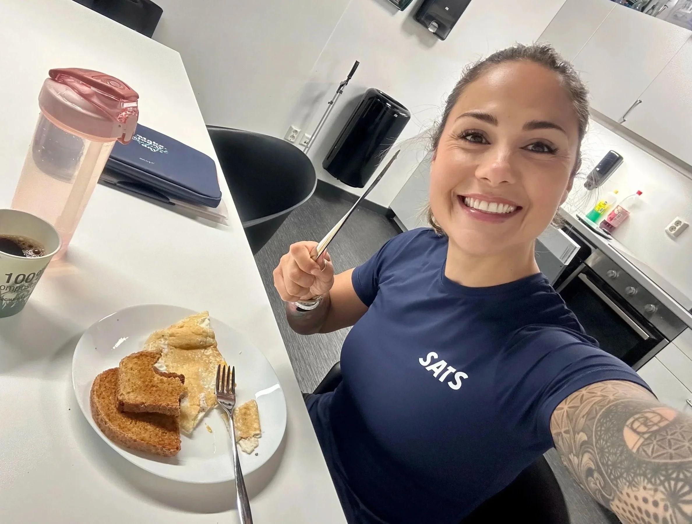 A smiling woman with a tattooed arm taking a selfie in an office break room. She is wearing a navy blue shirt with partially visible white text. The table in front of her has a plate with slices of bread, a cup of coffee, and a fork. There is a pink water bottle and a closed laptop on the table. The background features cabinets, an appliance, and cleaning supplies.
