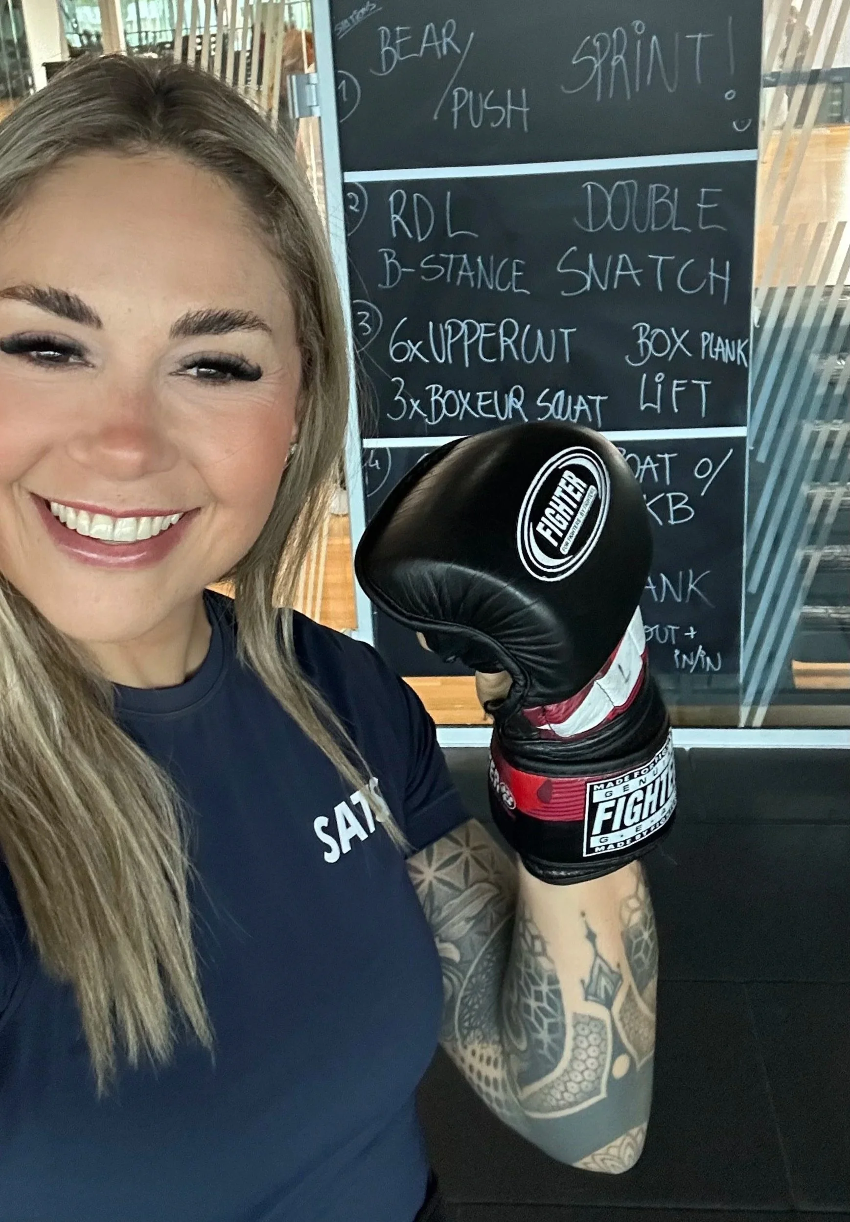 A smiling woman with long hair and tattoos on her arm, wearing a black shirt with white text, is holding a boxing glove with the fighter logo in a gym or boxing facility. Behind her, there is a blackboard with handwritten workout instructions.