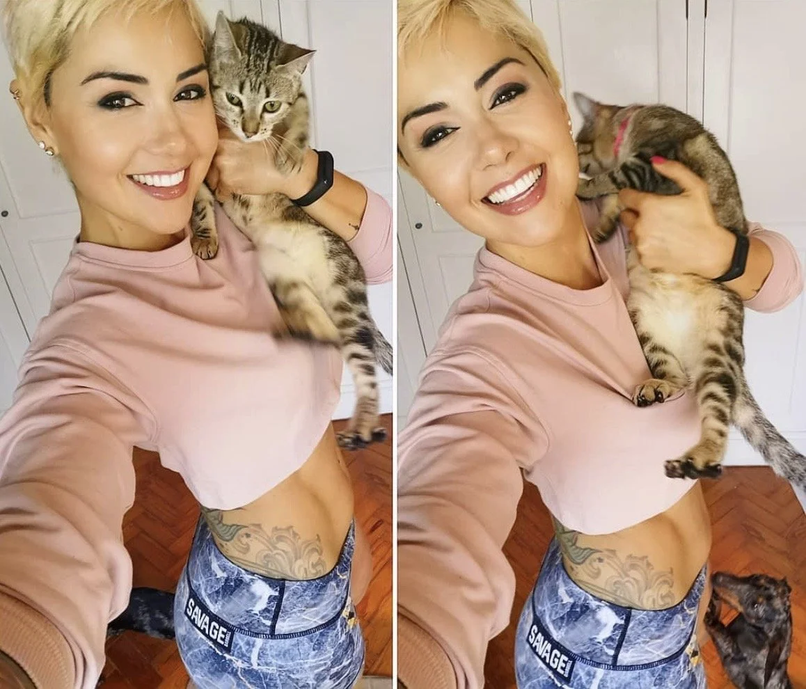 A smiling woman with short blonde hair, tattoos on her side and wearing a light pink long-sleeve shirt and blue camouflage leggings, holding a tabby cat in each hand, taking a selfie in front of a white door.