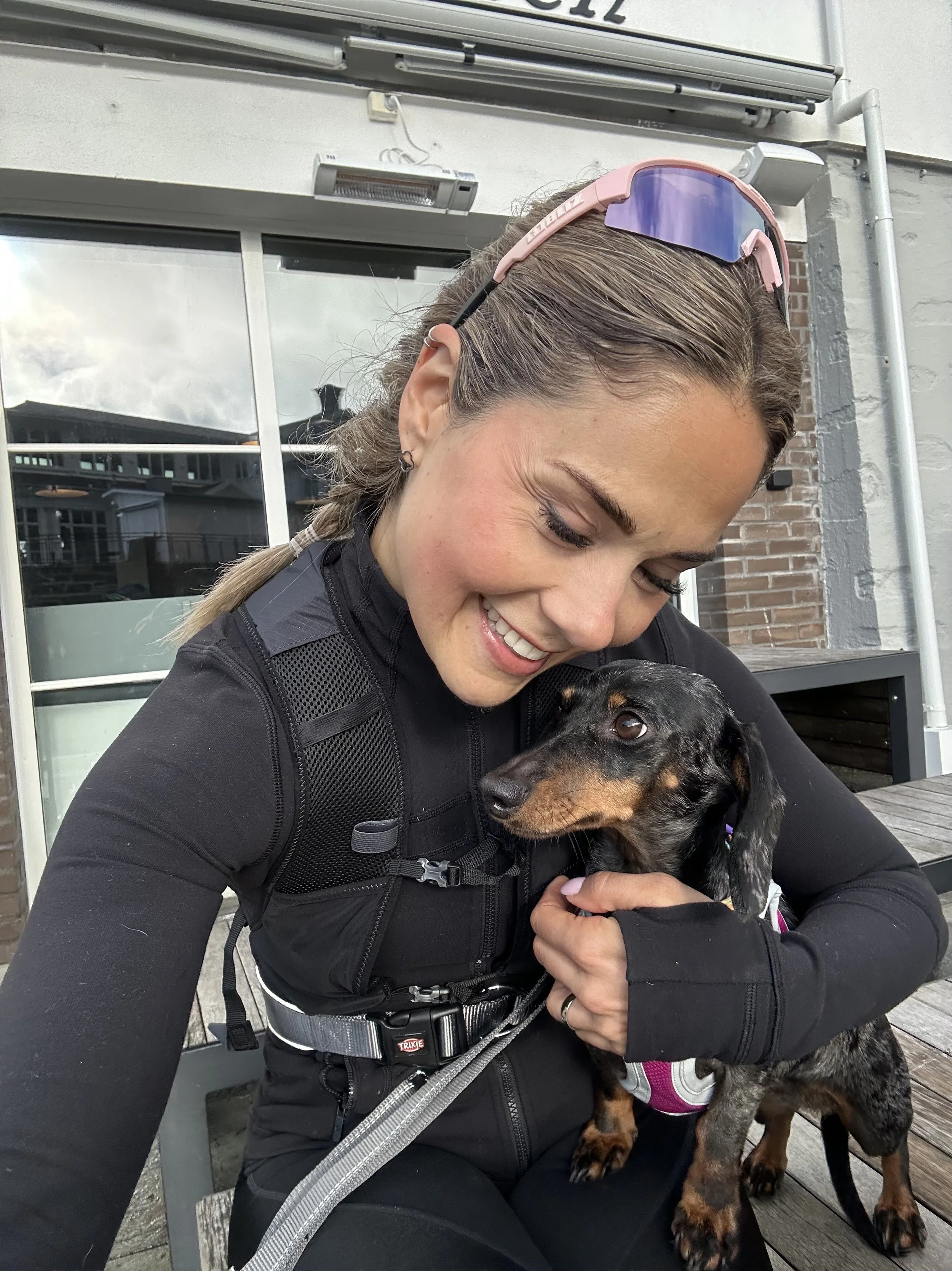 A woman with light brown hair in a braid, wearing pink sunglasses and a black jacket, smiling while holding a black and tan puppy wearing a pink vest, sitting on a bench outdoors.