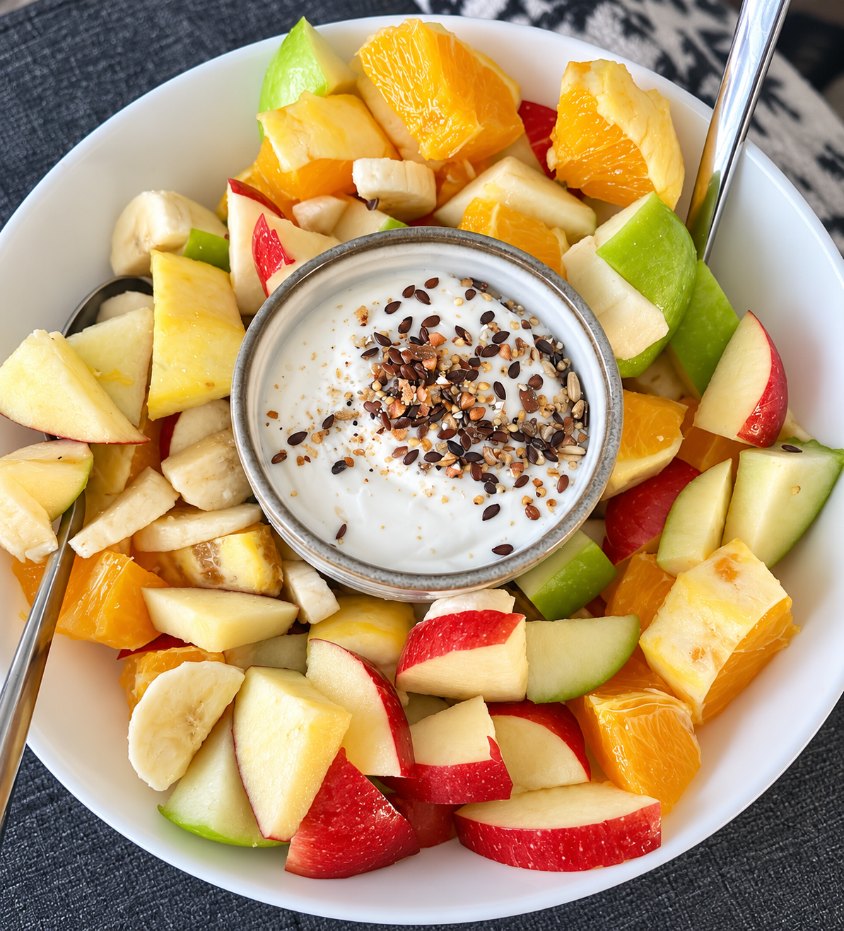 Bowl of mixed fresh fruit including apple, banana, orange, and melon pieces with yogurt topped with seeds in the center.