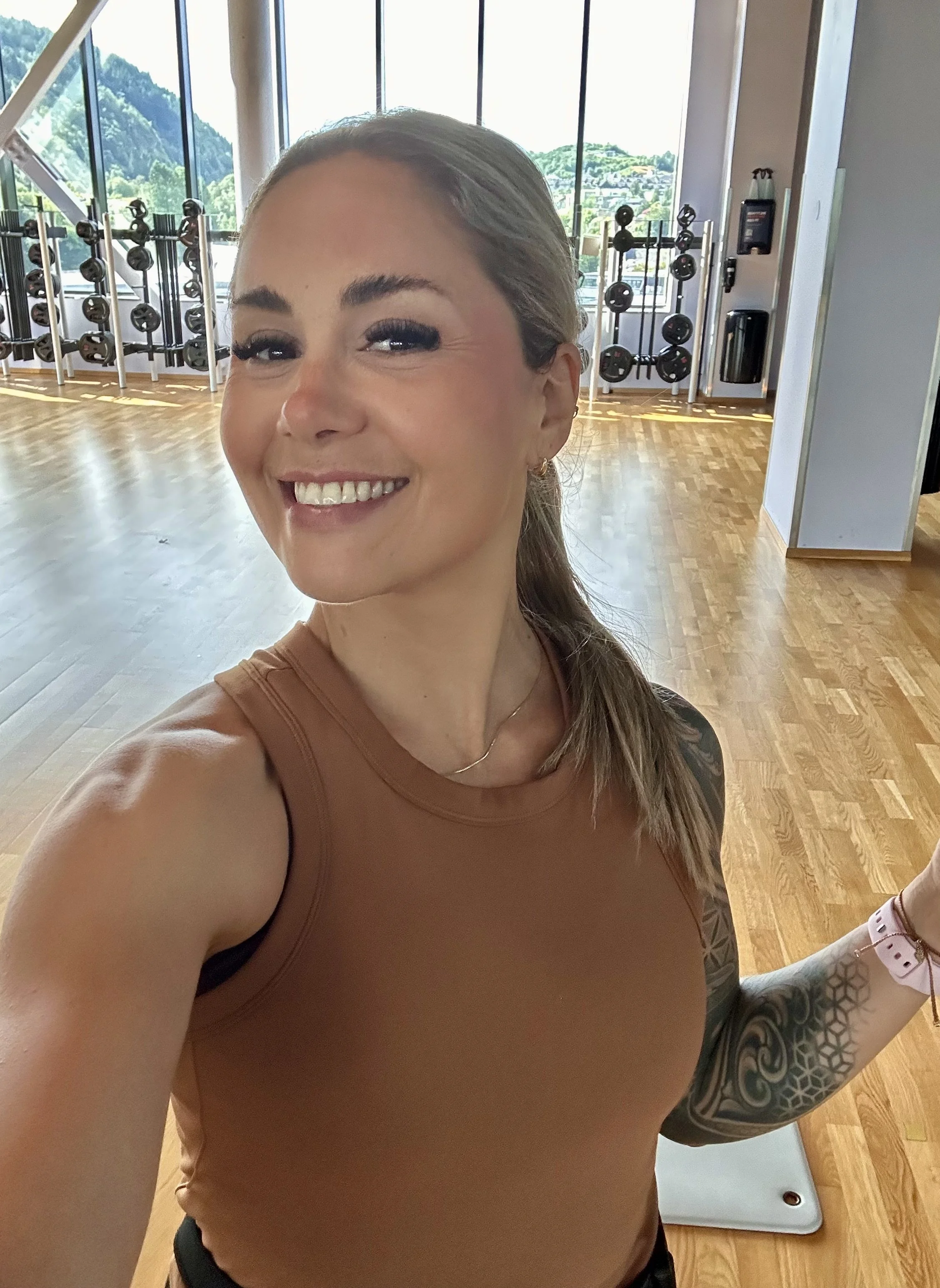 A woman taking a selfie in a gym, showing her smiling face, tattooed arm and shoulder, and workout attire, with gym equipment and large windows in the background.