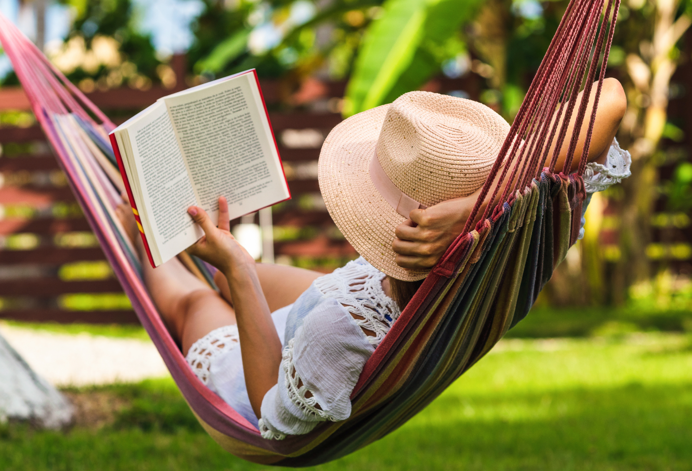 Person relaxing in a hammock outdoors, reading a book and holding a wide-brimmed straw hat.