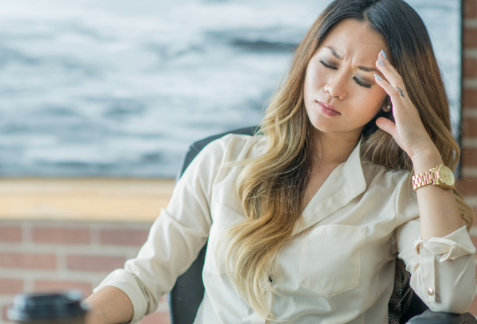 A woman sits at a desk, holding her forehead with a pained expression, with a body of water visible behind her.