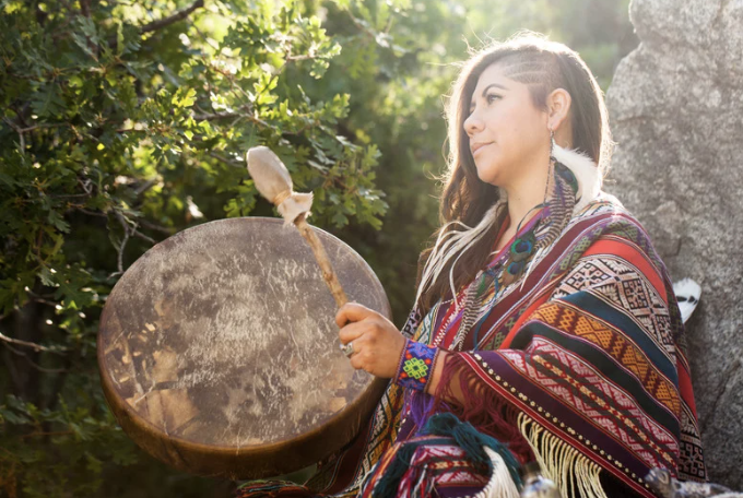 A young woman with long hair playing a drum outdoors, surrounded by green foliage, wearing a colorful, patterned shawl.