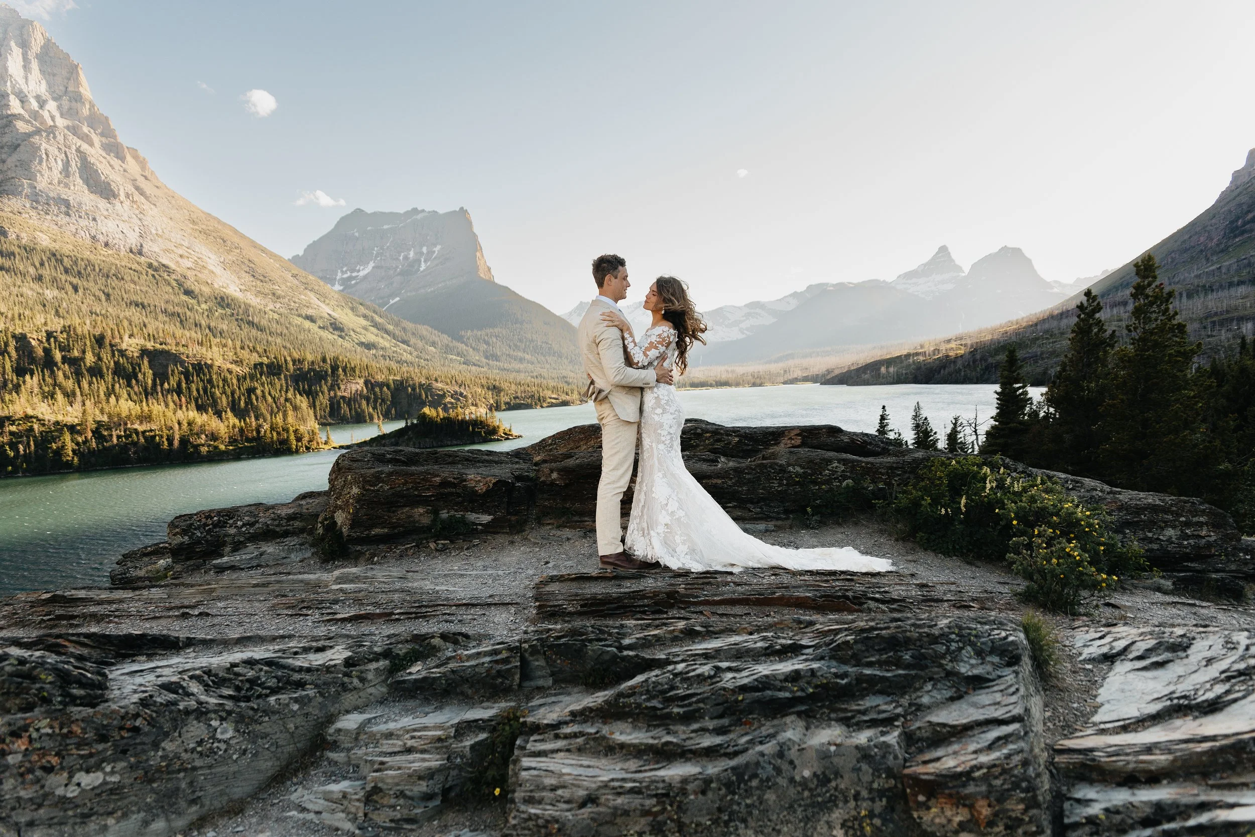 Glacier National Park wedding photography: bride and groom standing on rocks near a lake with mountains in the background, captured by a Montana photographer.