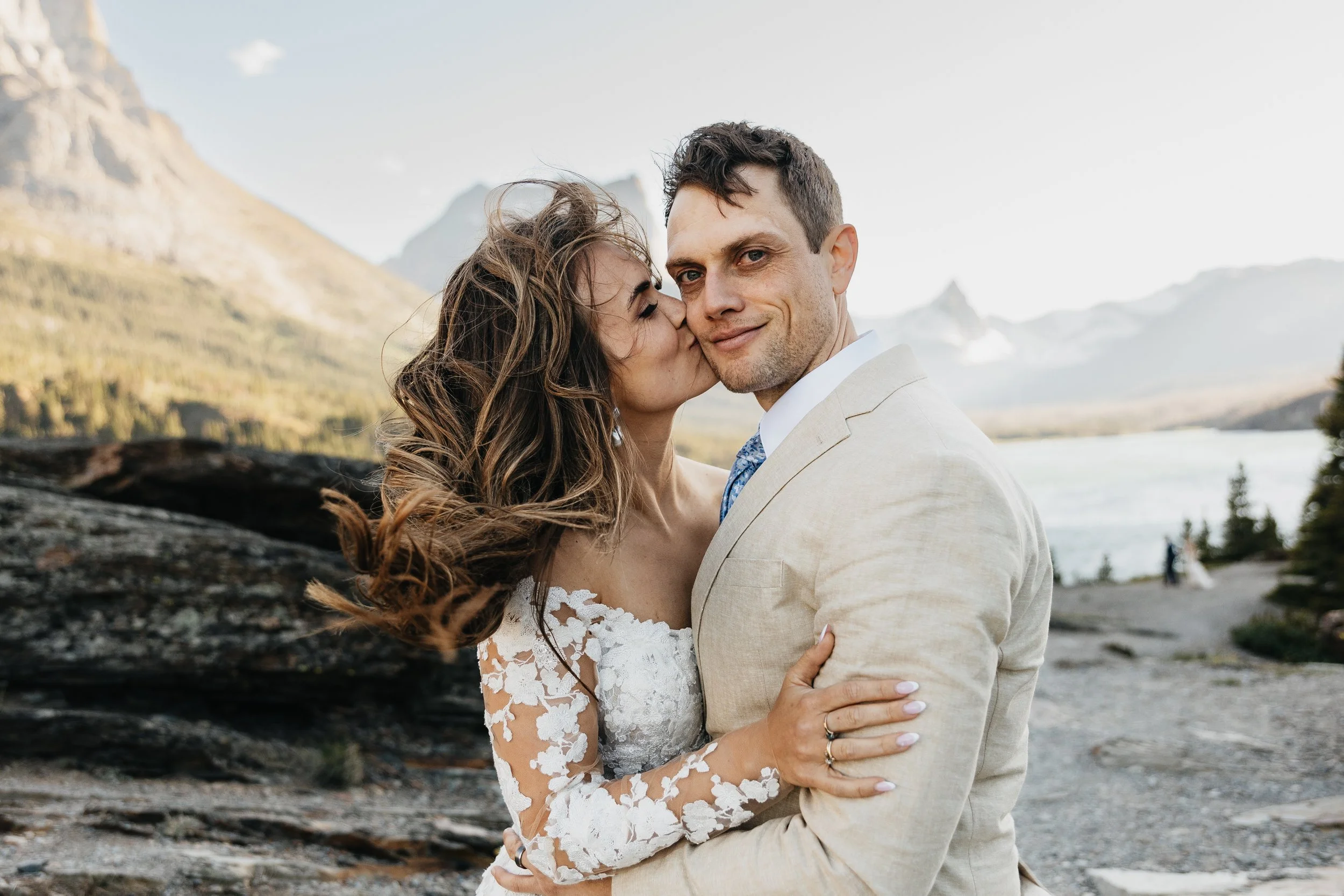 Missoula and Montana wedding photographer capturing a bride and groom embracing outdoors in Glacier National Park, with one kissing the other’s cheek against a mountain and lake landscape.