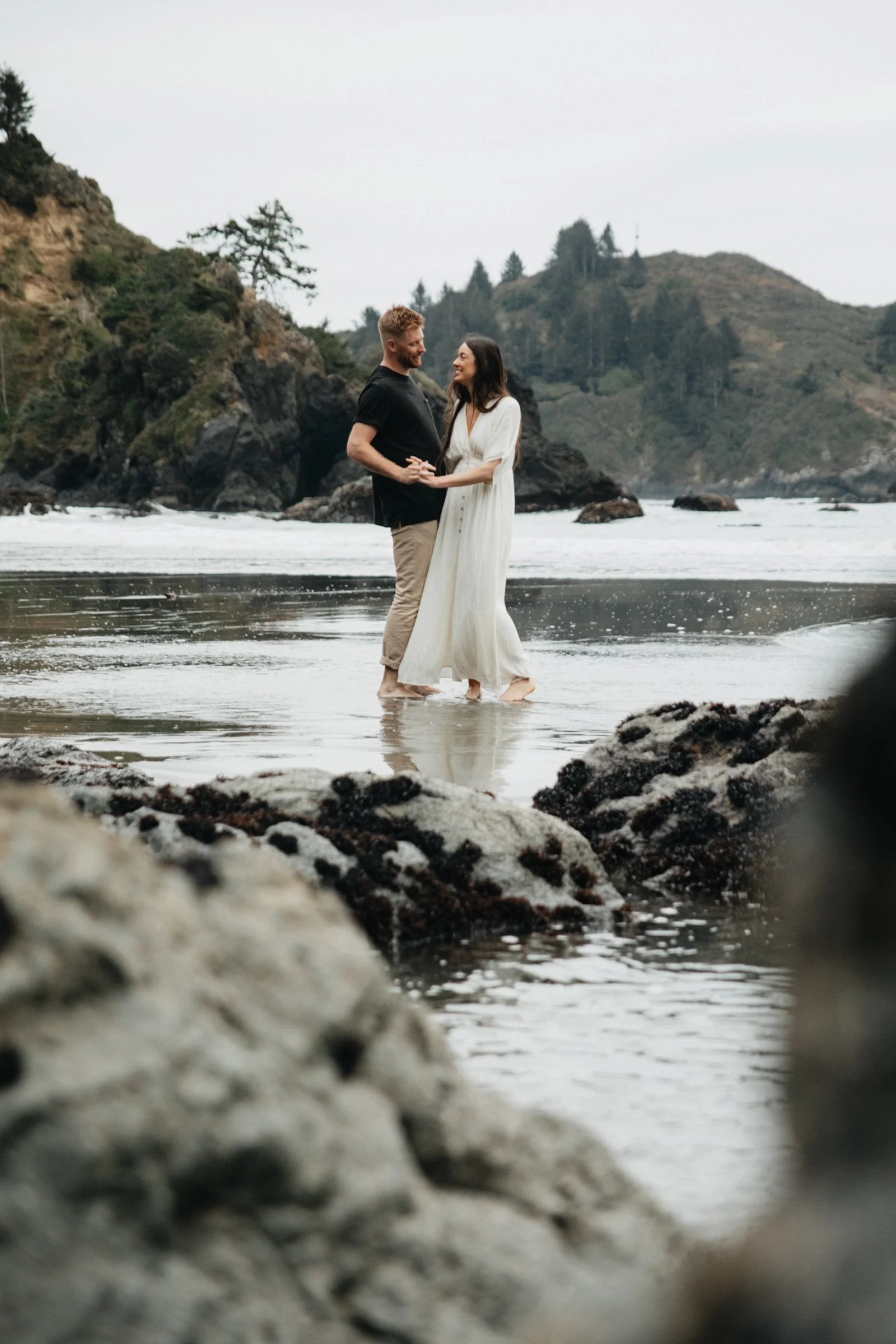 Engagement session on the Oregon Coast beach capturing candid moments between a couple near rocky cliffs and crashing waves.