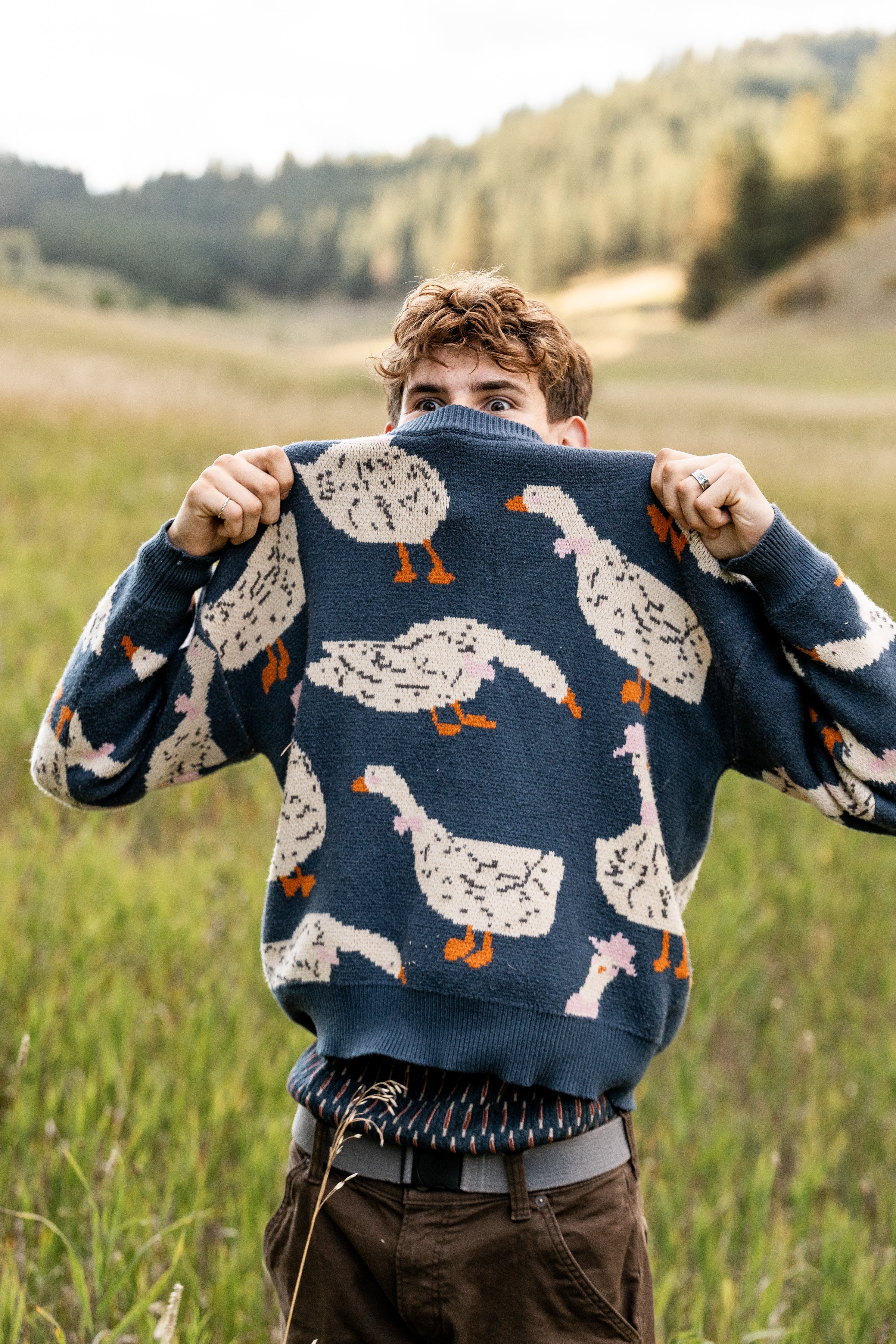 Person standing in a grassy field holding a sweater with a duck pattern over their face, with hills and trees in the background.