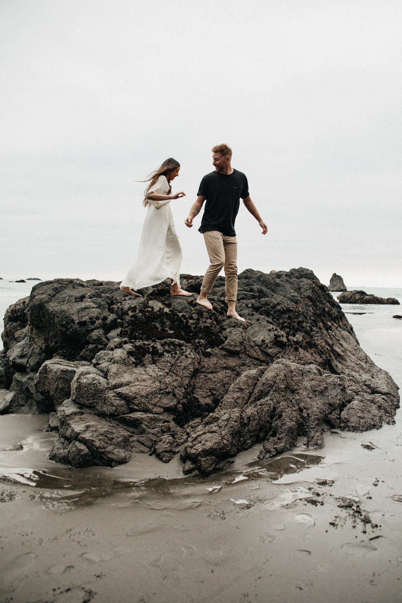 Natural engagement photography on the Oregon Coast with a couple standing by the ocean, showcasing movement, connection, and coastal scenery.