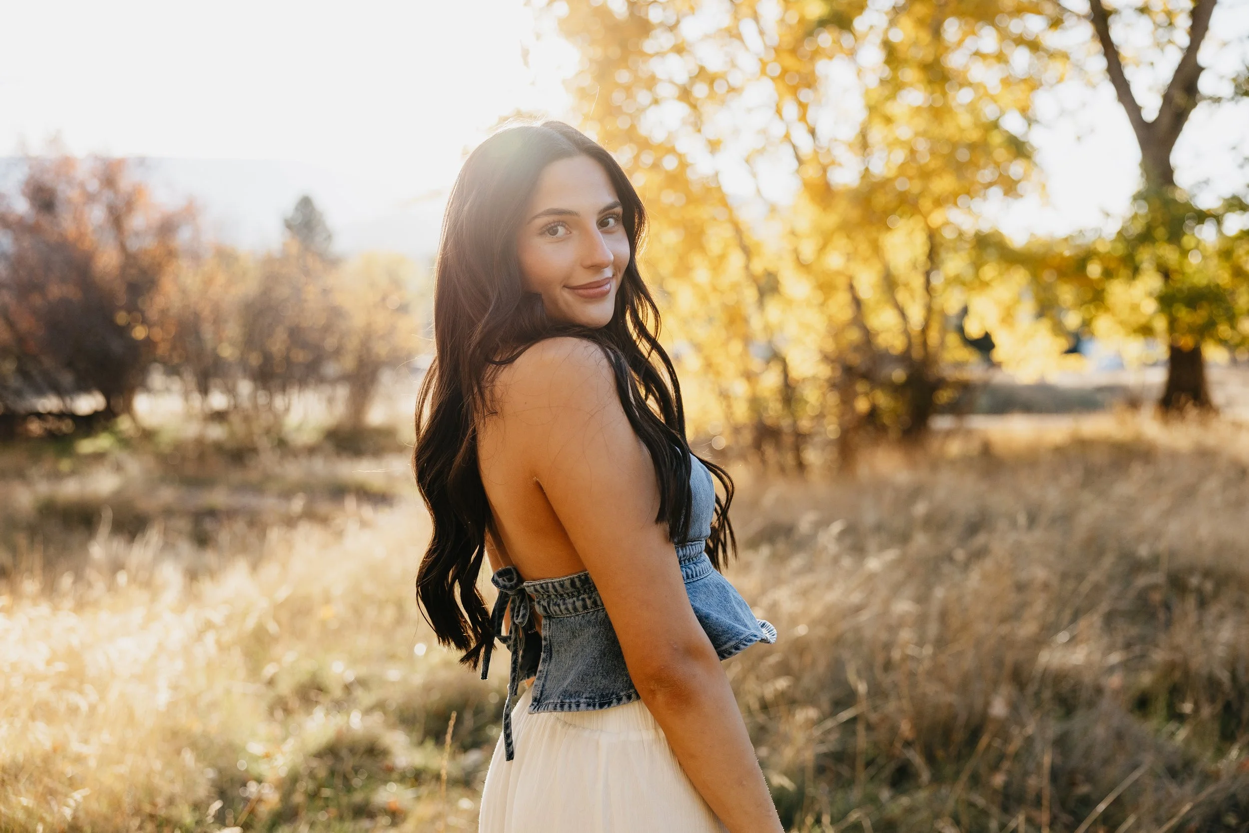 A young woman with long dark hair standing outdoors in a field with trees showing fall colors, illuminated by the warm sunlight.