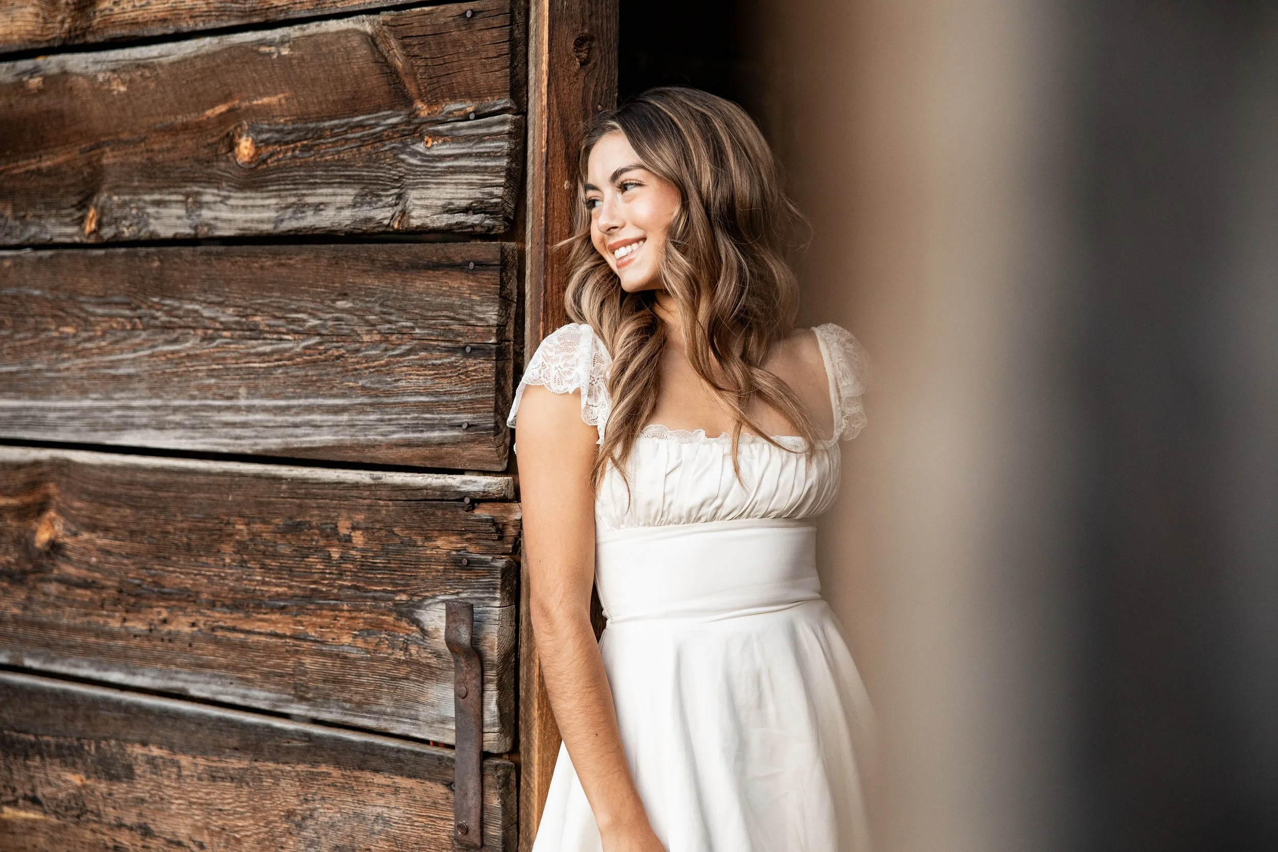 A young woman with wavy brown hair smiles as she leans against a rustic wooden wall, wearing a white dress with lace details on the sleeves.
