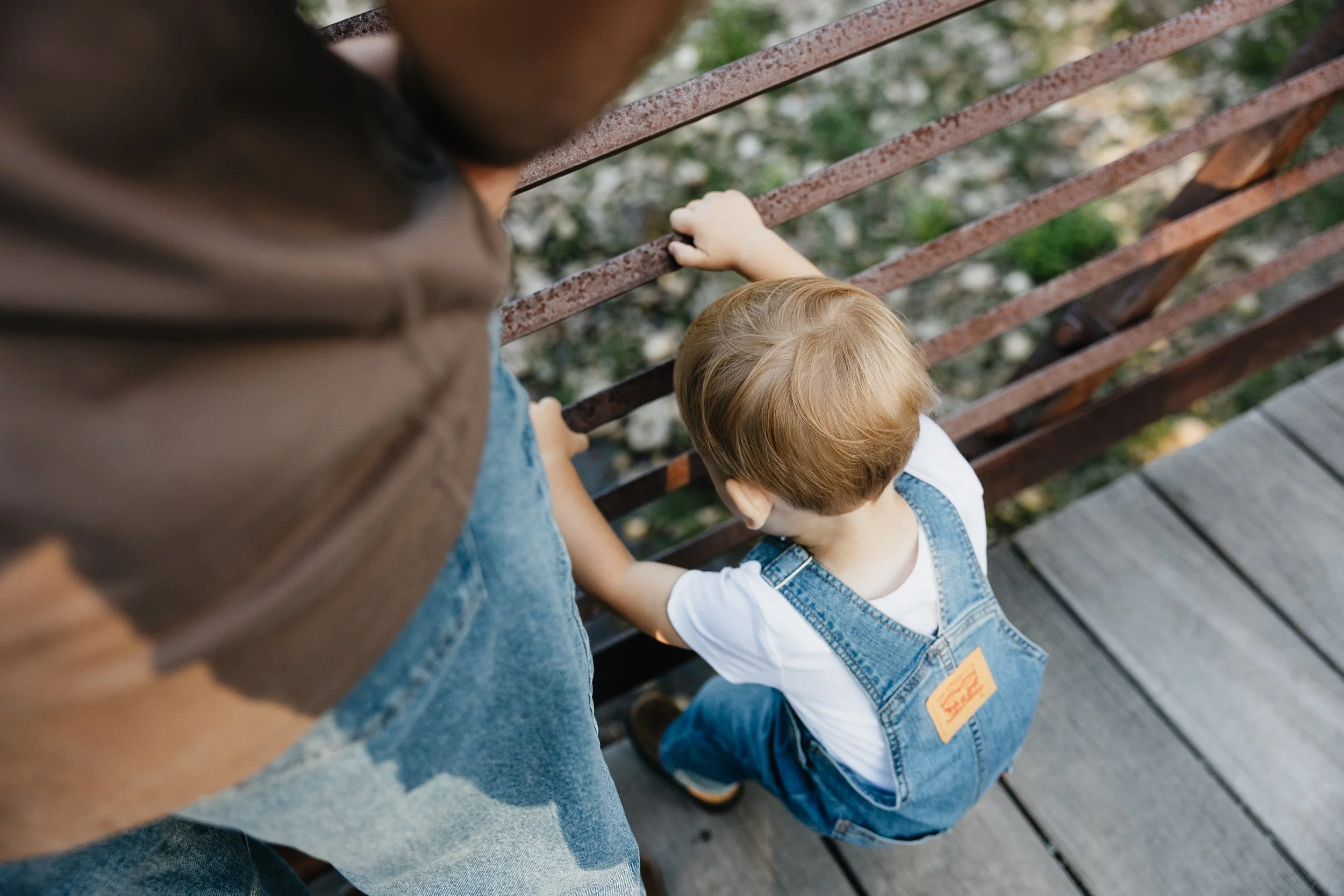 A young boy with red hair in denim overalls holding onto a rusty metal railing on a wooden bridge, seen from above.