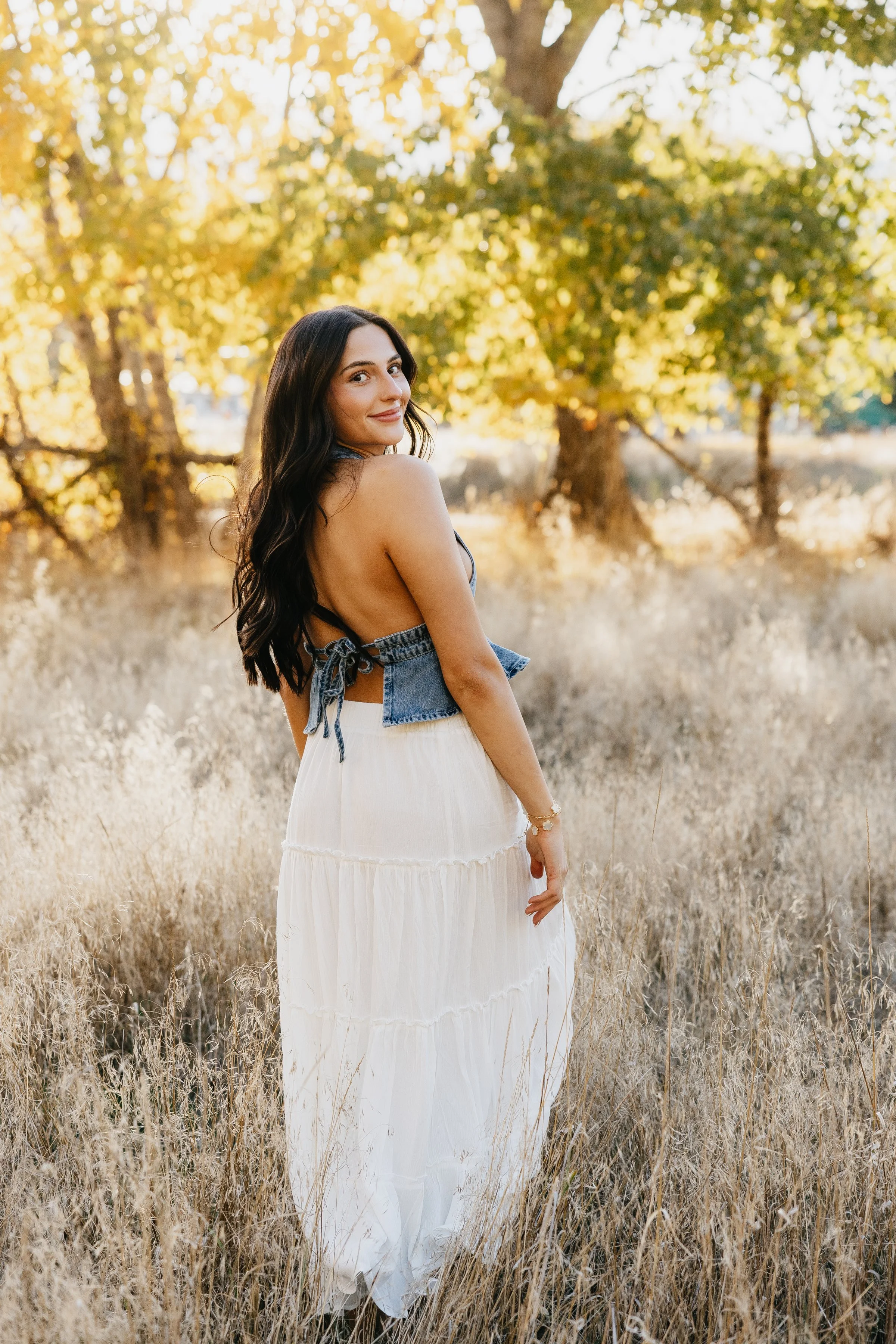 A woman with long dark hair in a white maxi skirt and denim top standing in a golden field with trees behind her, during sunset.