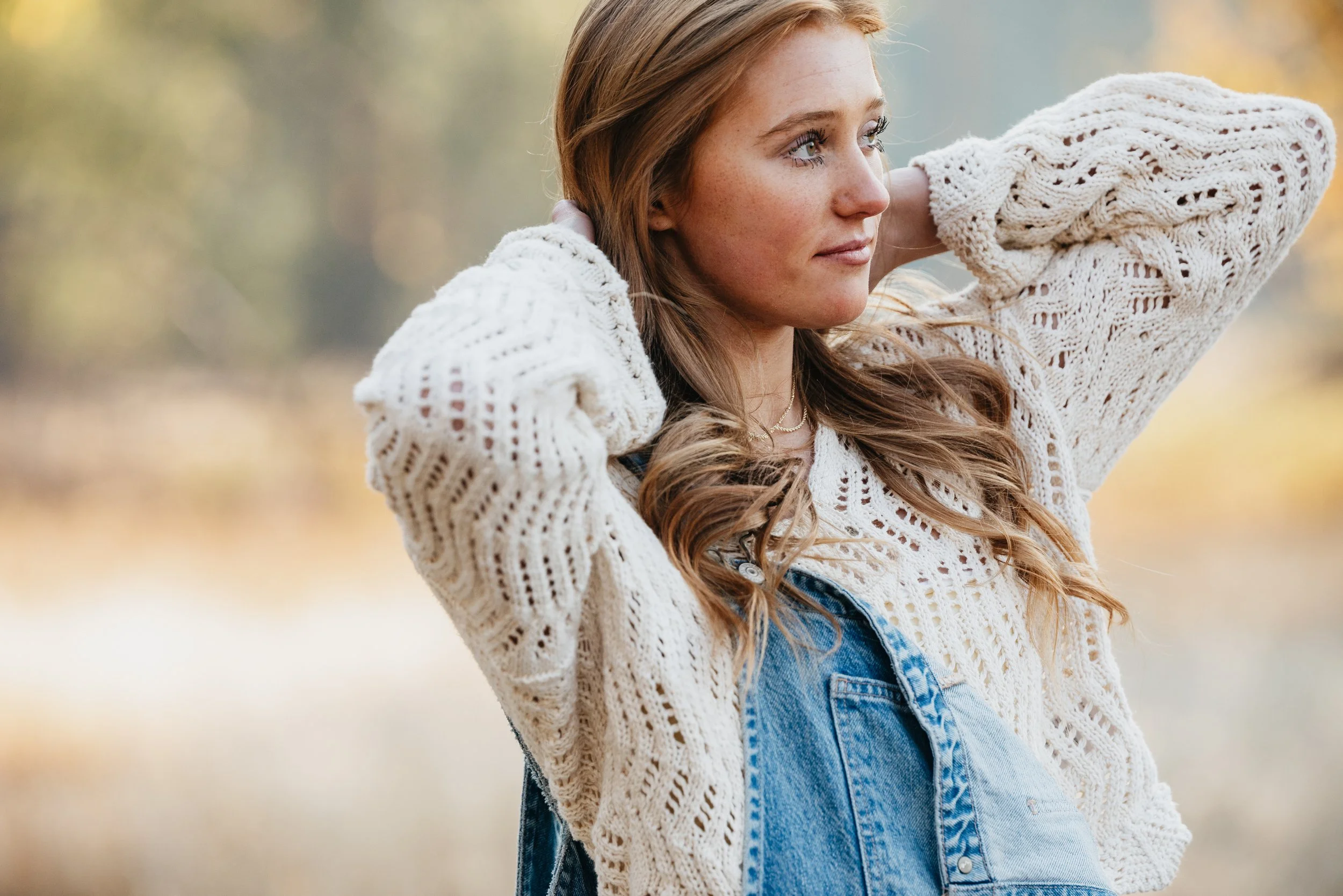 Young woman with long wavy hair, wearing a knitted sweater and denim overalls, standing outdoors in a natural setting with autumn foliage, her hands behind her head looking to the side.