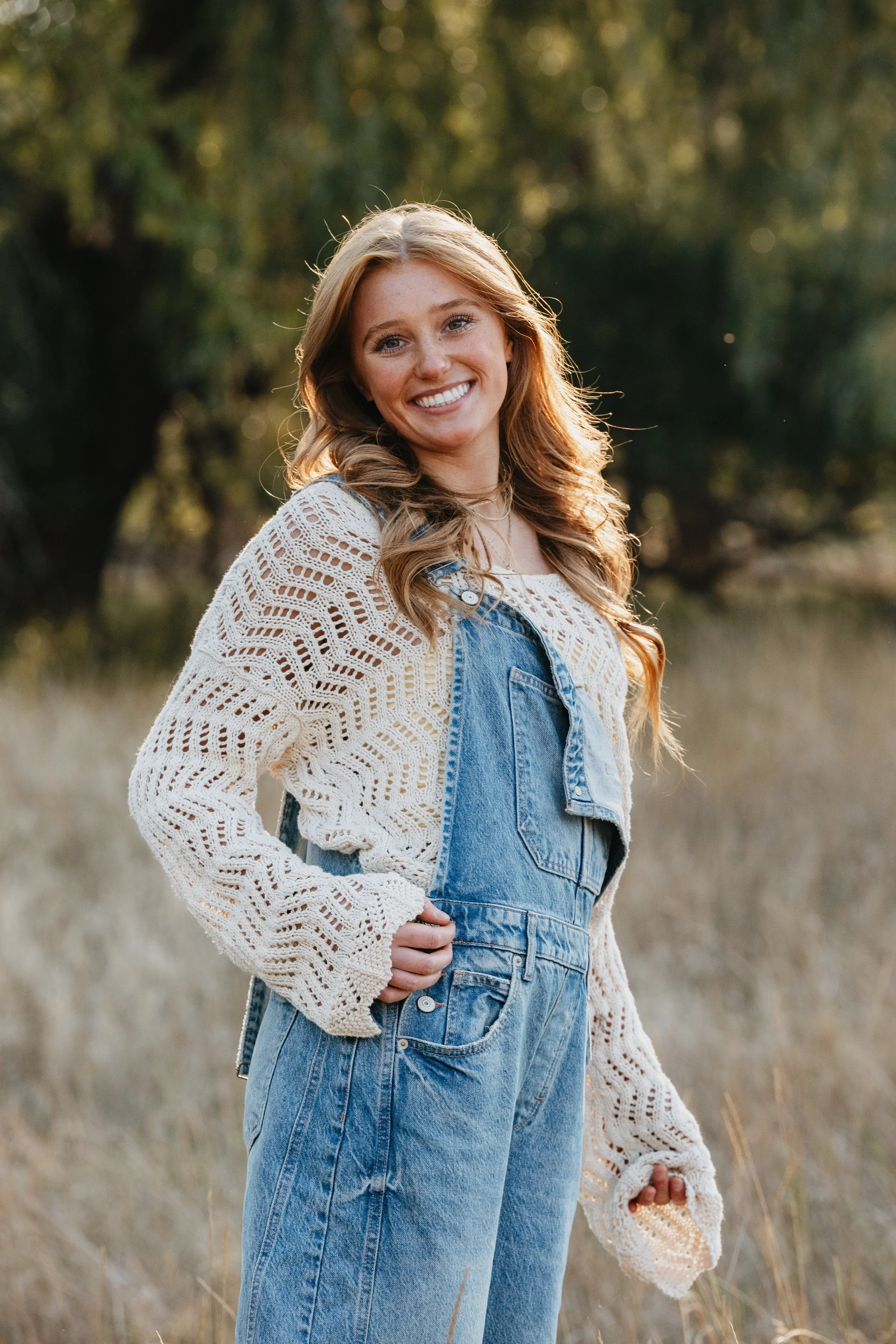 A young woman with long, wavy blonde hair and a bright smile standing outdoors in a field with trees in the background, wearing a white crochet sweater and denim overalls.