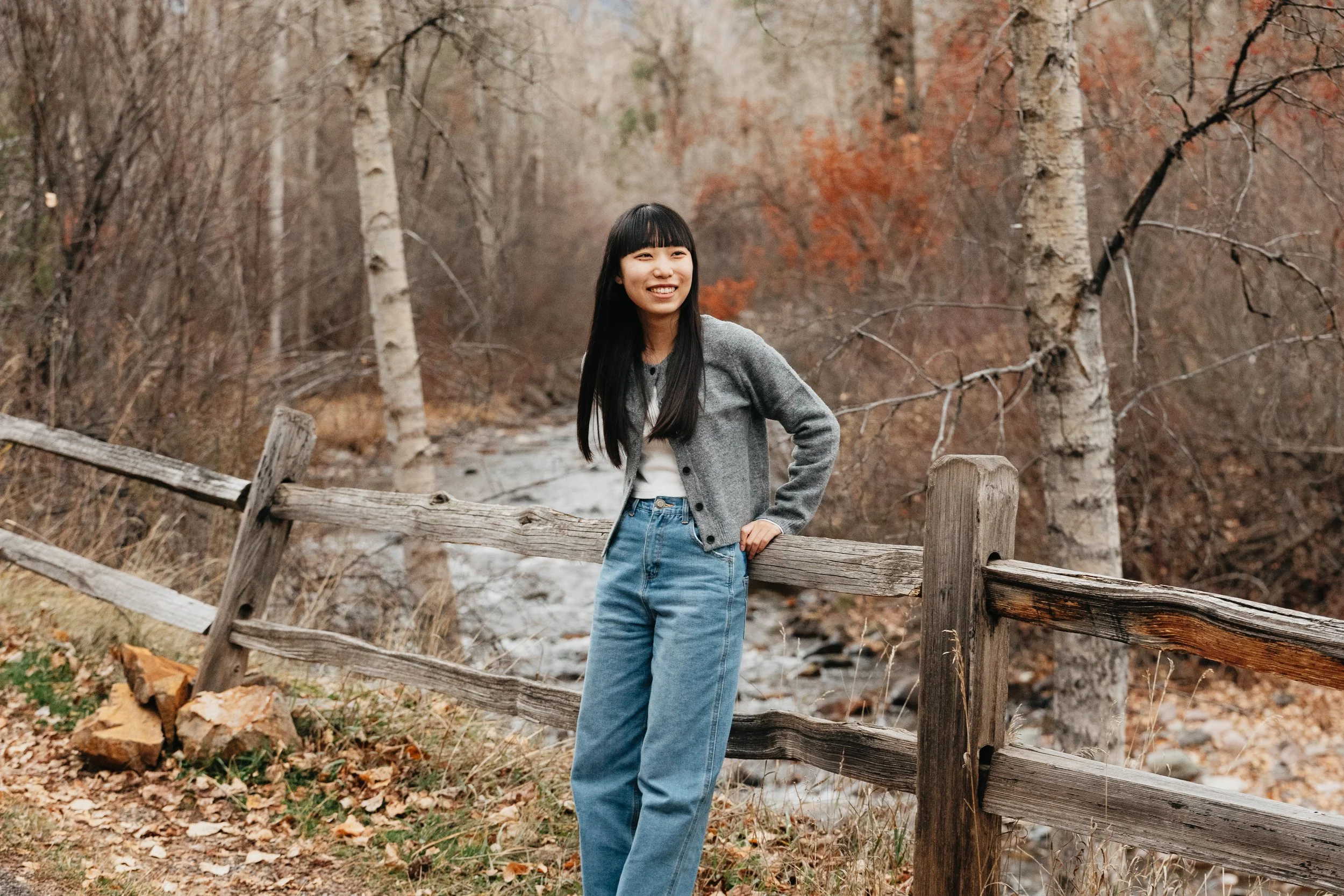 A smiling young woman with long black hair, wearing a grey cardigan and blue jeans, standing by a wooden fence along a creek in a forest with autumn-colored trees.