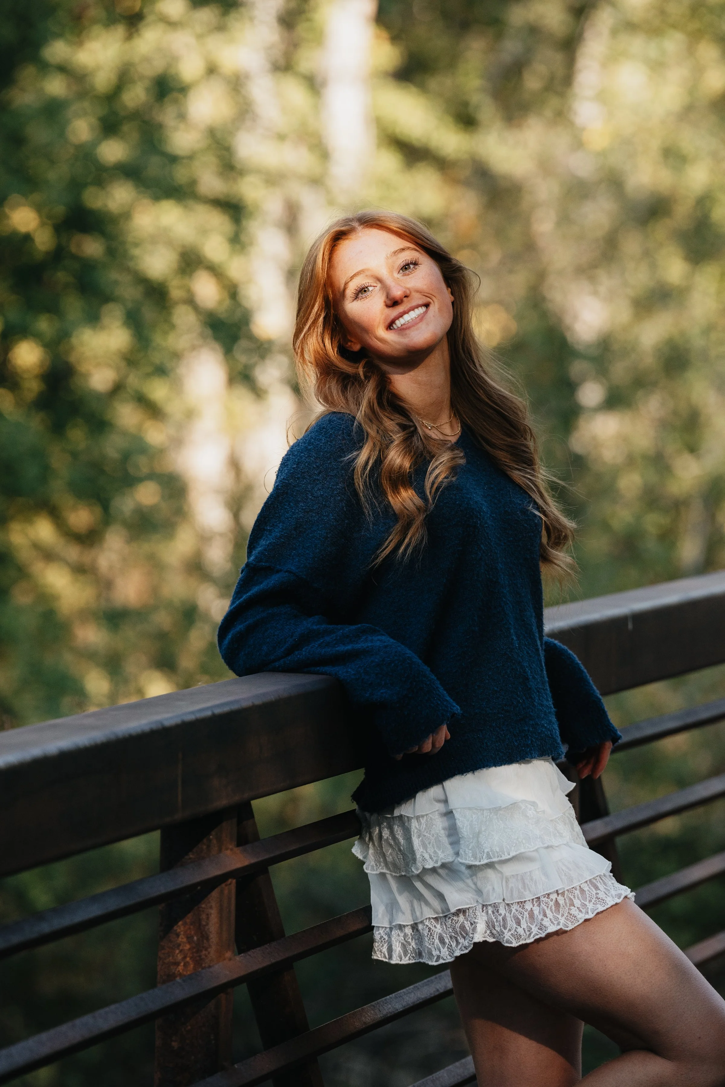 Smiling young woman with long wavy hair wearing a blue sweater and white lace skirt, standing outdoors in a park, leaning on a wooden railing, with blurred trees in the background.