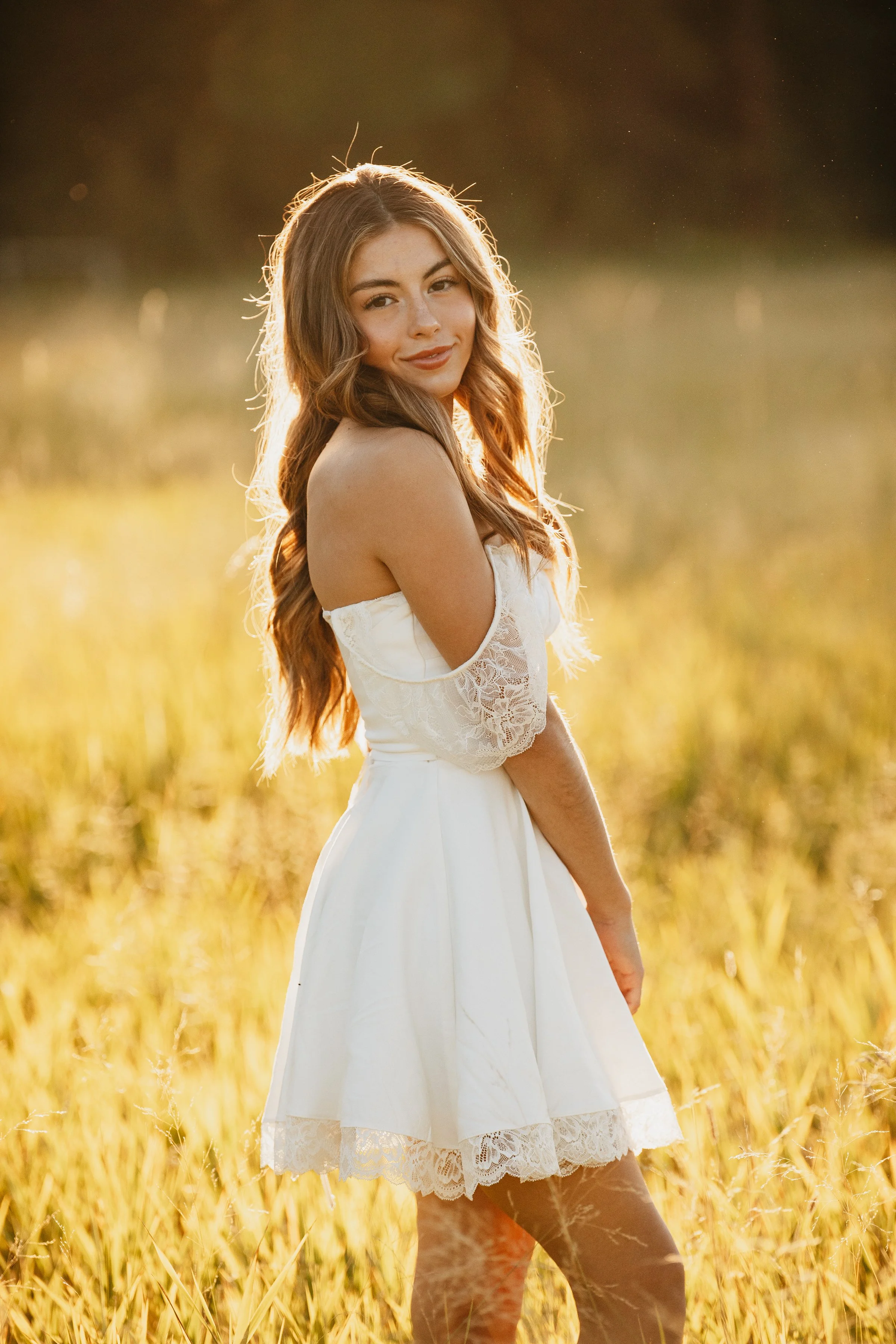 A young woman with long wavy hair standing in a field of tall grass during sunset, wearing a white dress with lace sleeves.