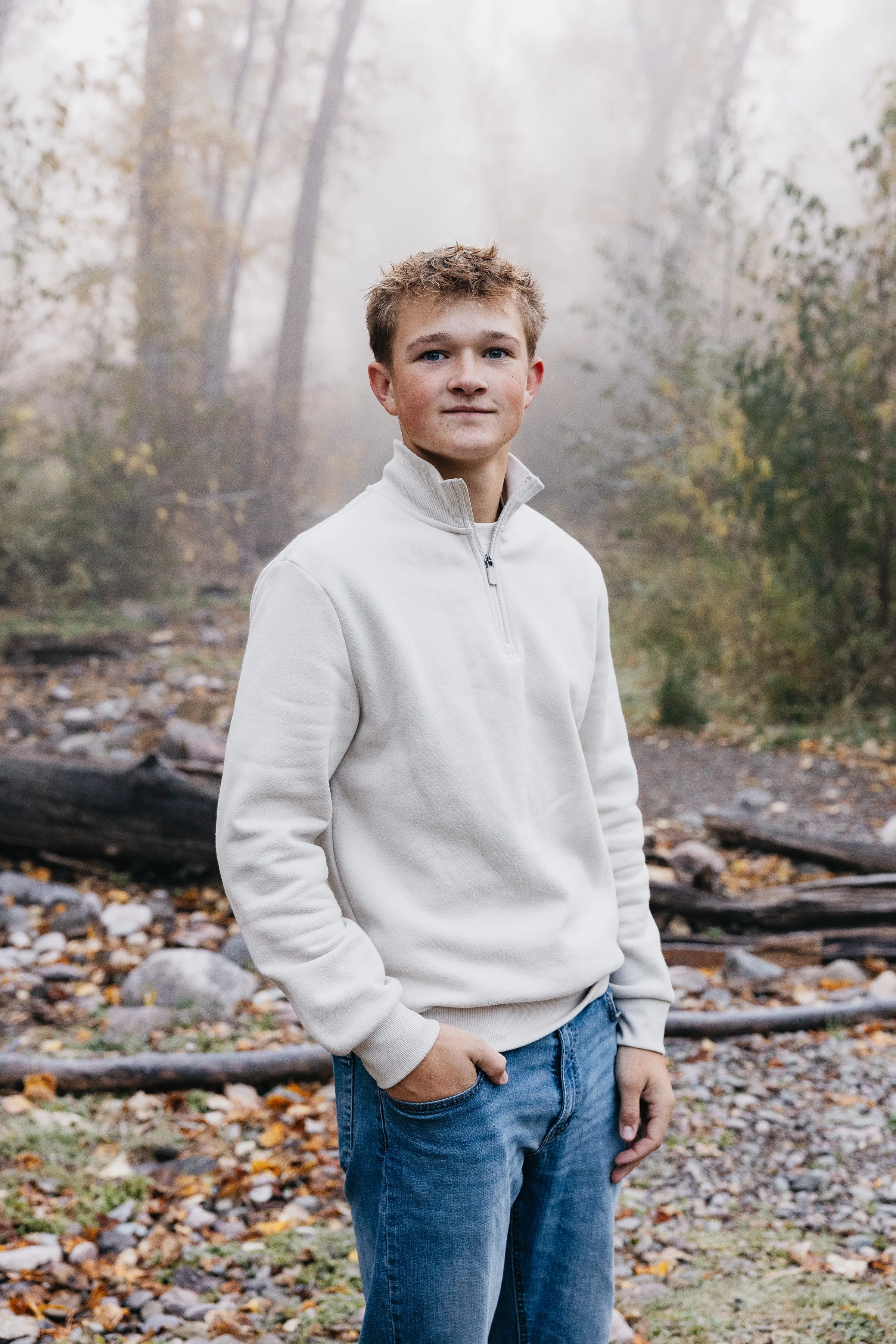 A young man with light brown hair and blue eyes wearing a light gray quarter-zip sweater and blue jeans standing outdoors in a foggy, wooded area with fallen autumn leaves and logs on the ground.