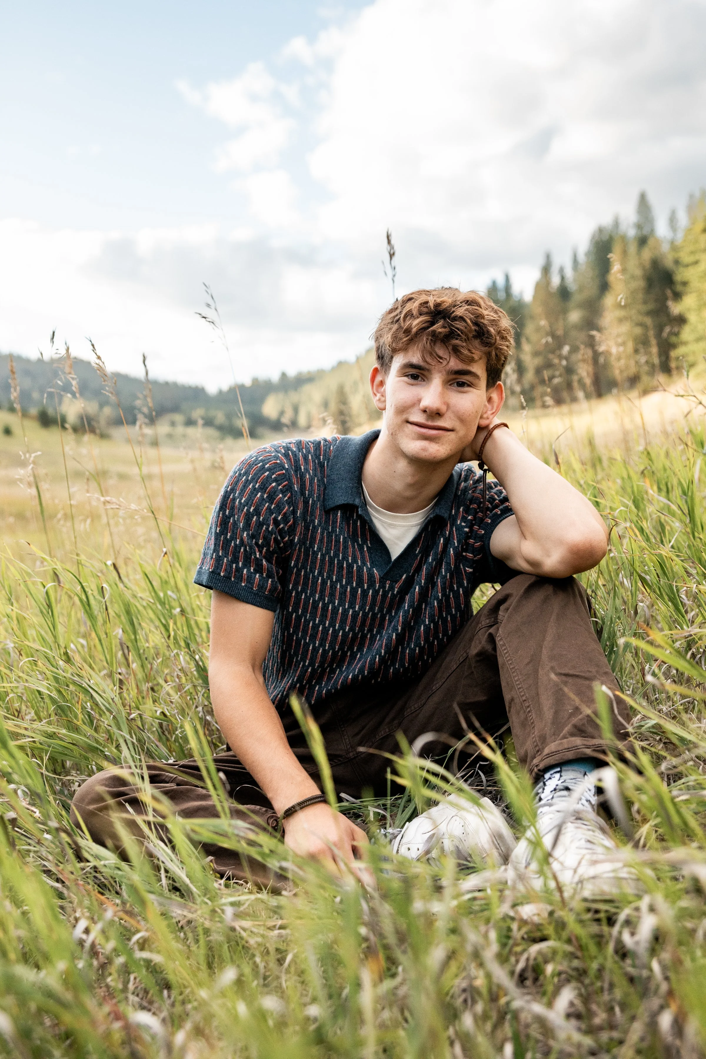 A young man sitting in tall grass in a field, with trees and hills in the background under a partly cloudy sky.