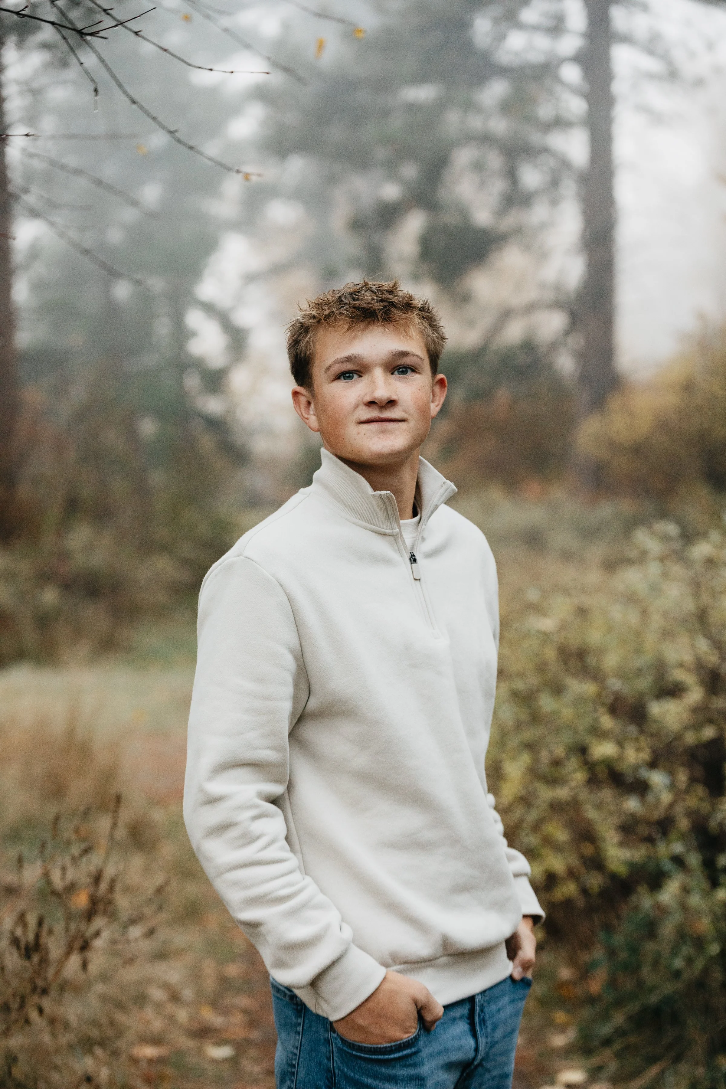 A teenage boy with short, light brown hair, wearing a cream-colored quarter-zip pullover and jeans, stands outdoors in a forested area with fog and autumn foliage in the background.