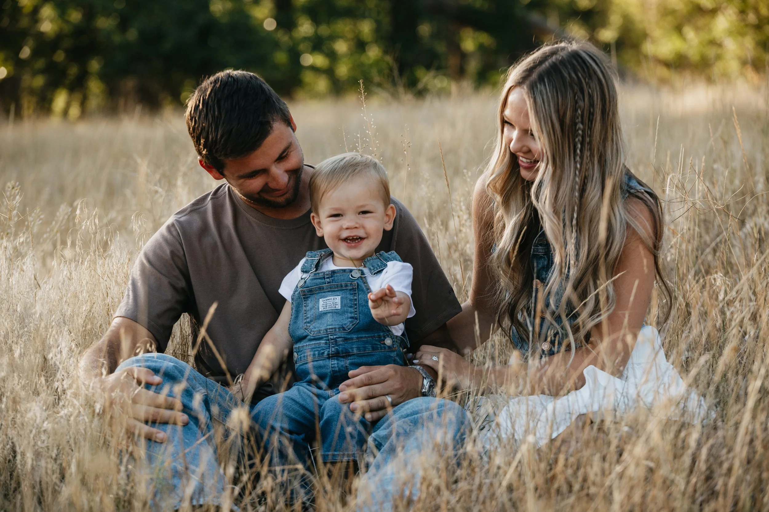 Family of three sitting in tall grass outdoors, smiling and laughing together.