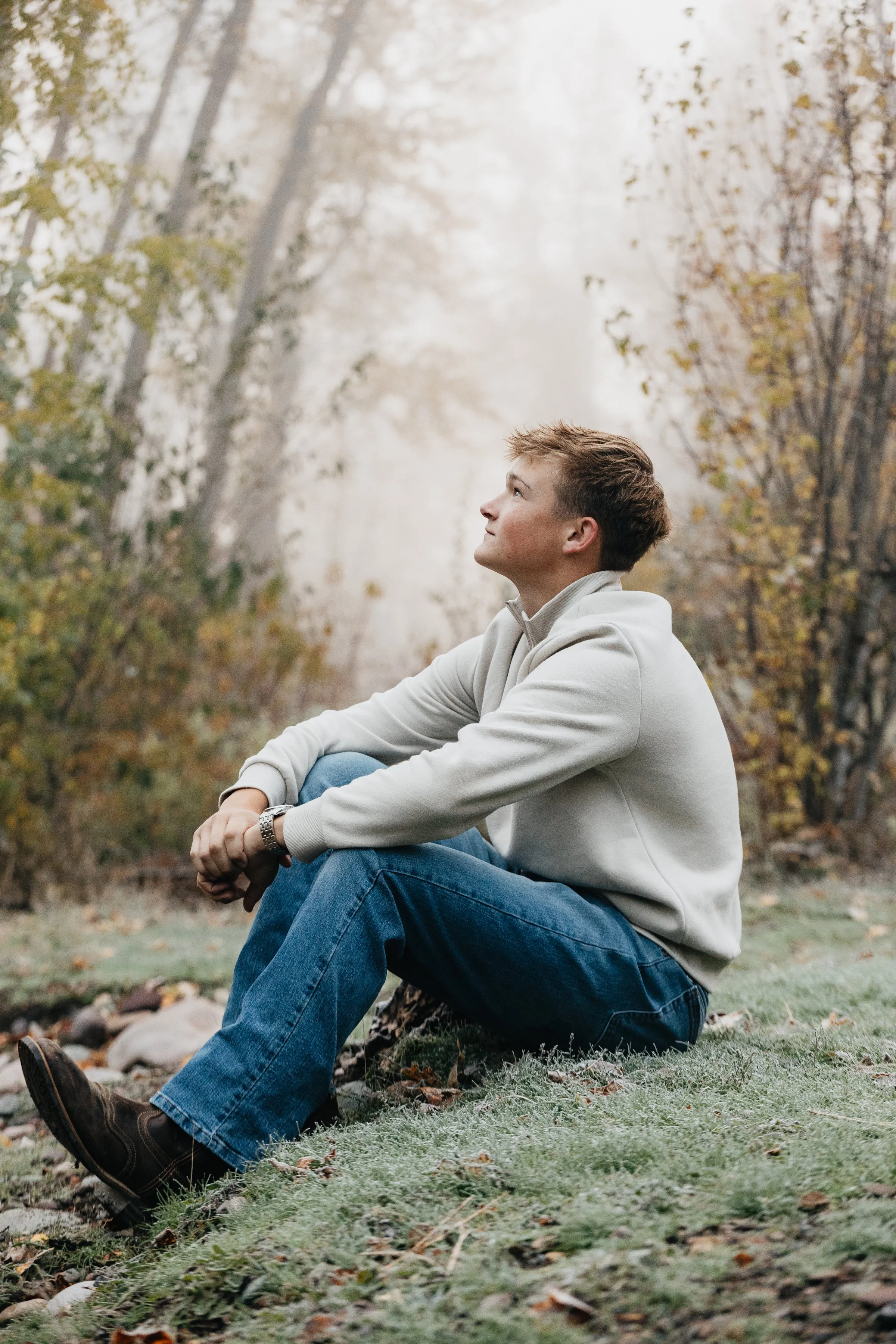 A young man sitting on the grass in a forested area during fall, looking up thoughtfully.