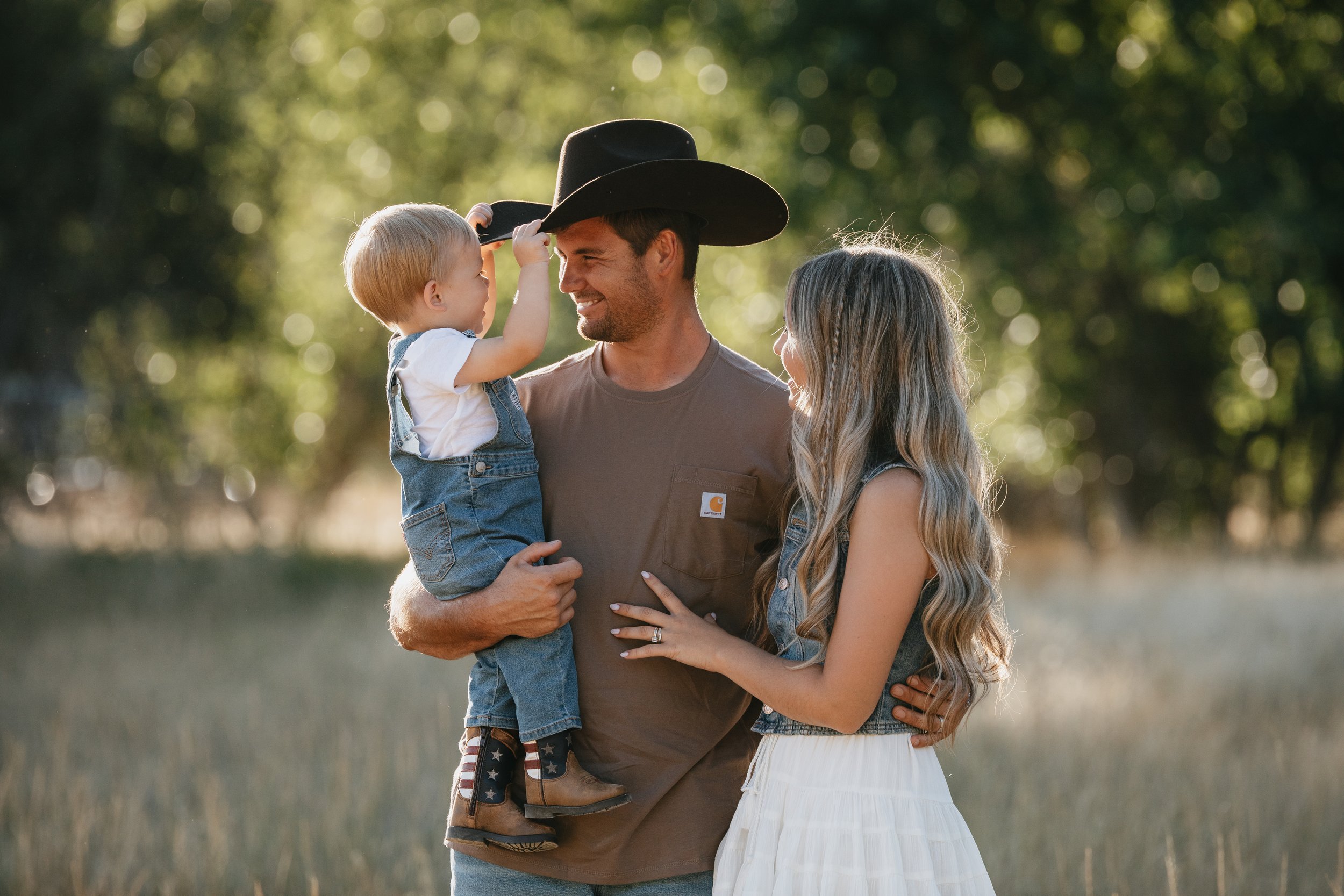 A family of three, father, mother, and young son, outdoors in a natural setting with green trees in the background. The father is holding the son, who is fitting a black cowboy hat on his head, while the mother looks on smiling. The father and son ar