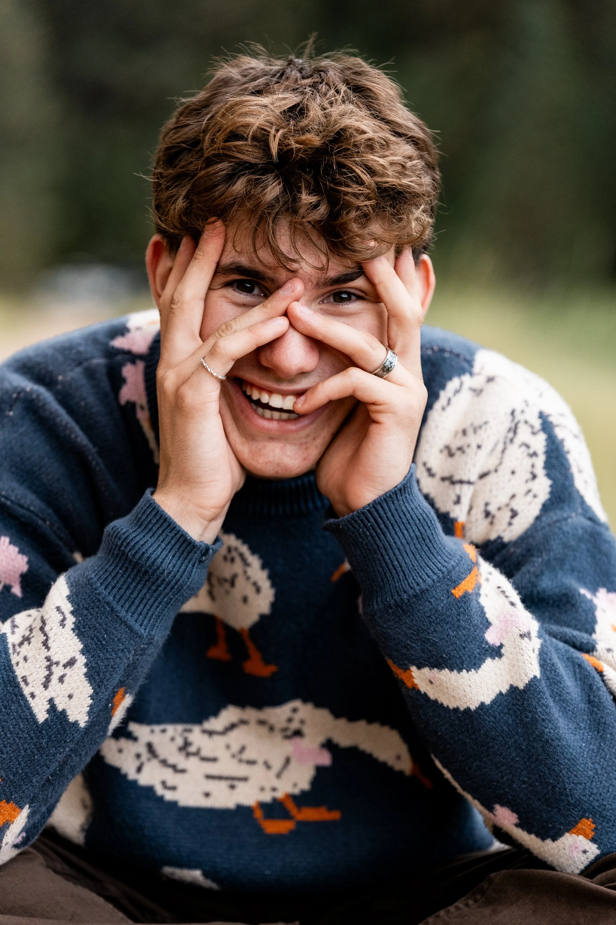 A young man with curly brown hair smiling and making a playful face, with his hands around his face, wearing a navy blue sweater with white and orange patterns, outdoors with blurred greenery in the background.