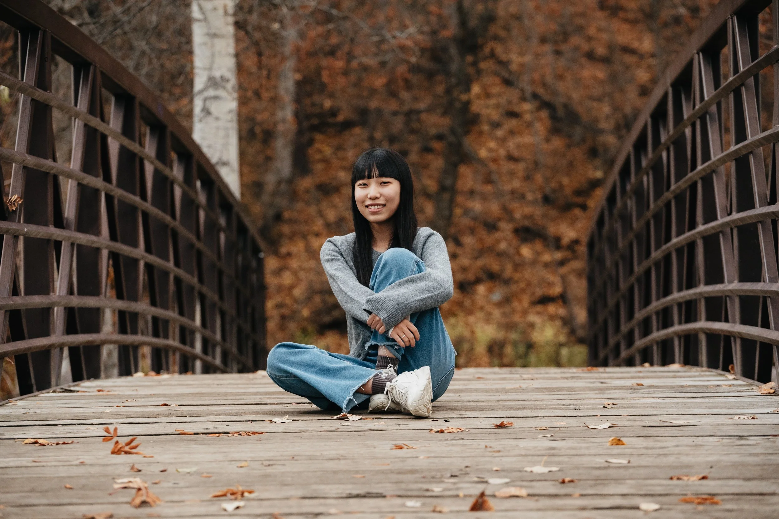 A young woman sitting cross-legged on a wooden bridge surrounded by autumn trees with colorful fall leaves.