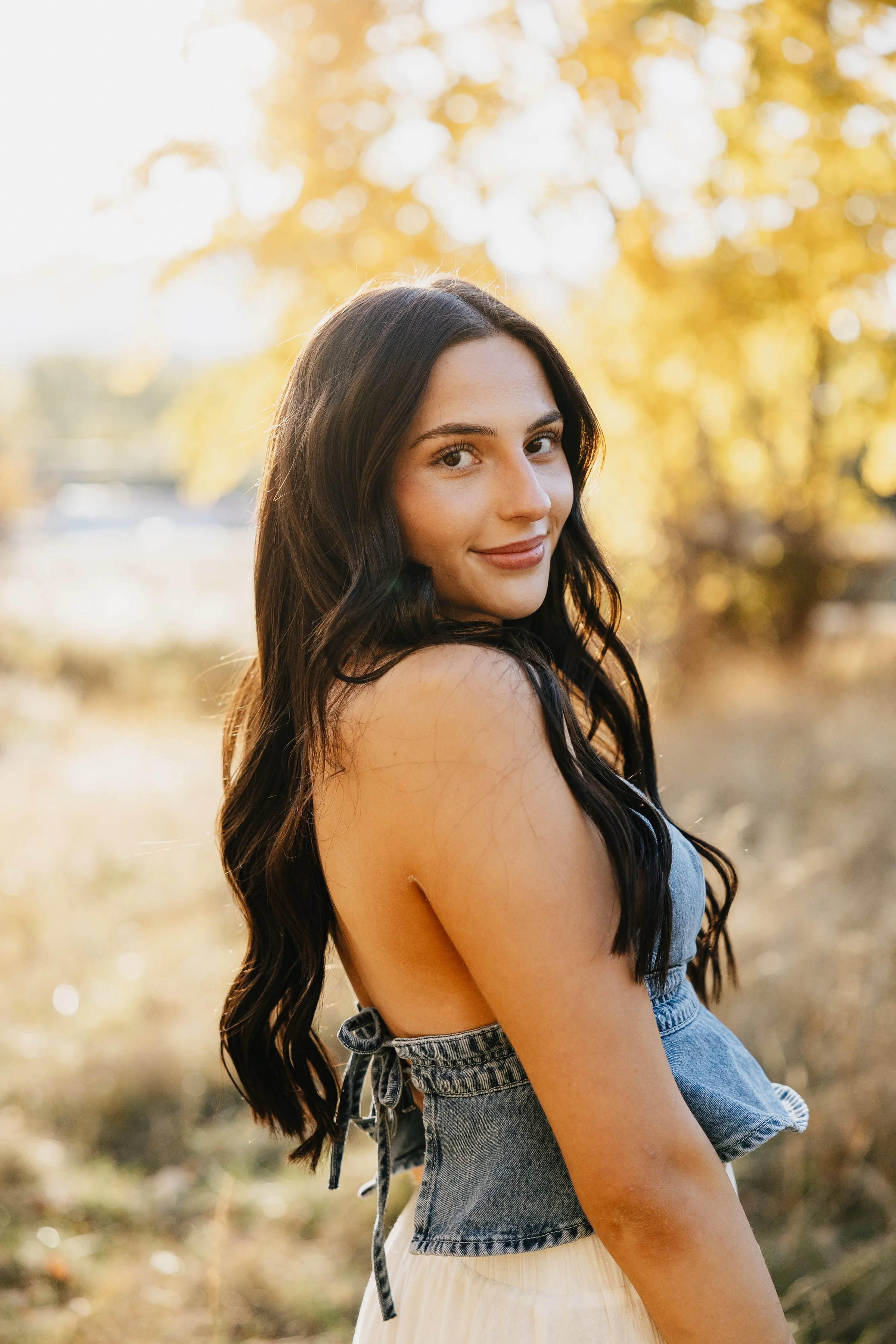 A young woman with long dark hair stands outdoors in sunlight, smiling softly. She is wearing a sleeveless denim top and a light-colored skirt with a blurred natural background of trees and foliage.
