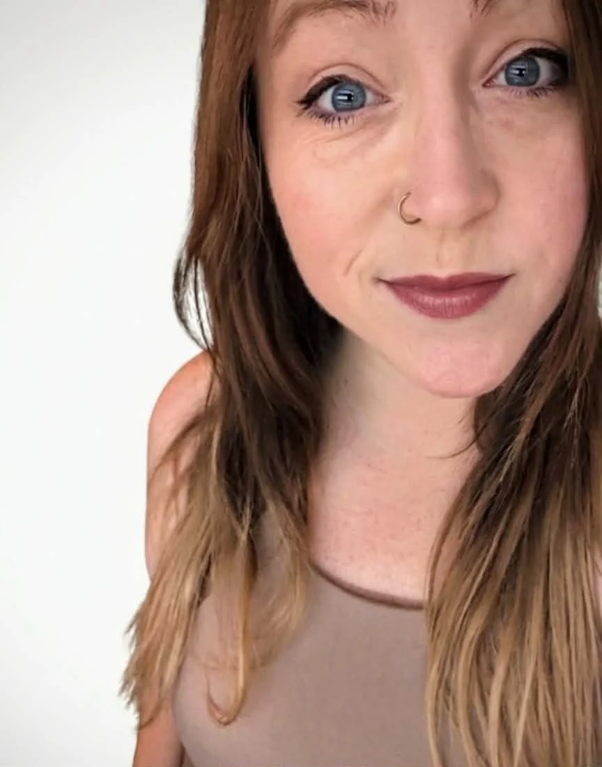 Close-up of a woman with blue eyes, red hair, winged eyeliner, a nose ring, and wearing a beige top.