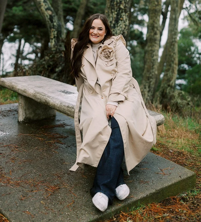 Smiling woman sitting on a stone park bench outdoors, surrounded by trees, wearing a beige trench coat with a flower decoration, dark jeans, and white sneakers.