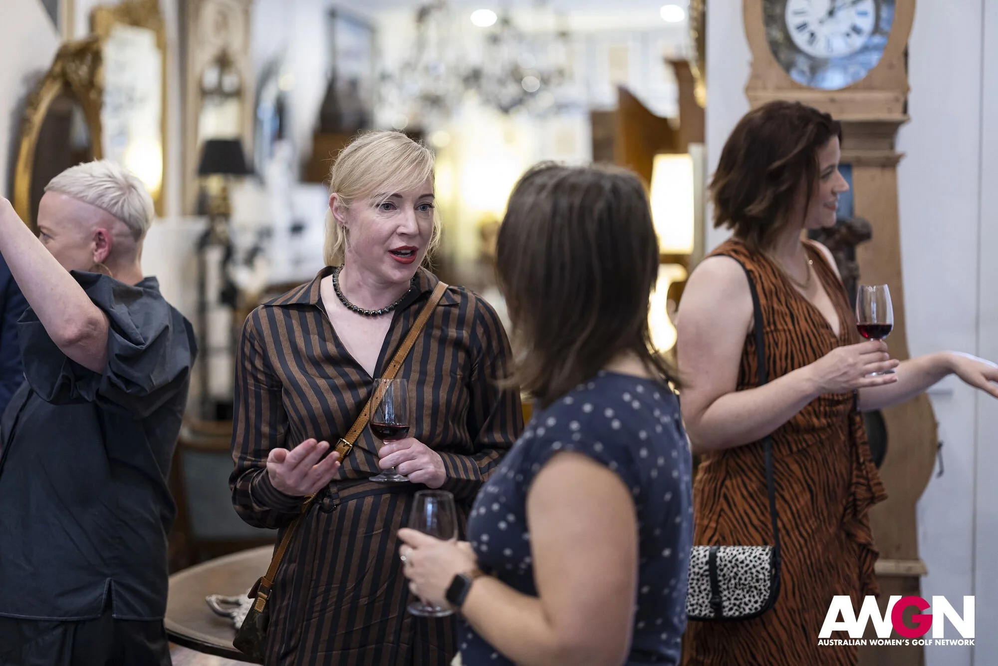 Group of people at a social event, holding wine glasses and talking in an indoor setting with decorative items and a clock in the background. AWGN logo visible.