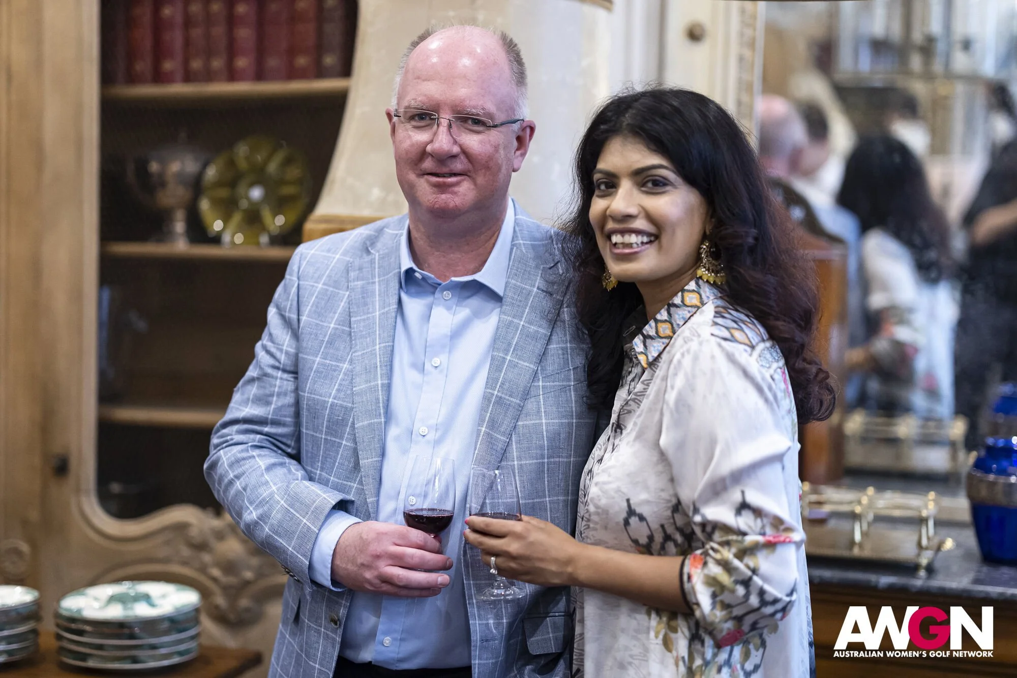 A man in a blue suit with glasses and a woman with long dark hair holding wine glasses, smiling in a decorated room with a mirror reflection and the AWGN logo in the corner.