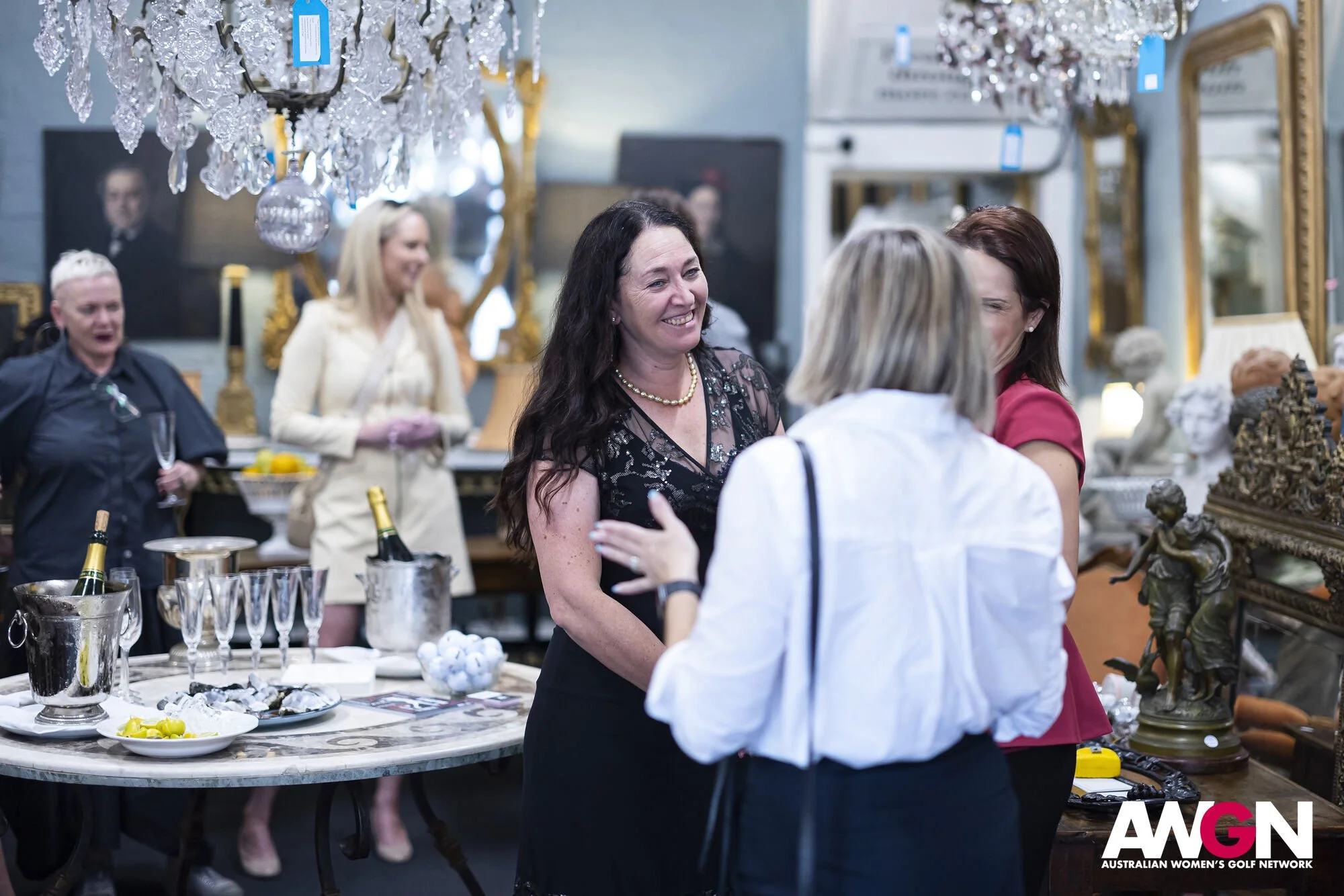 People socializing in a decorative room with chandeliers and antique decor, including champagne bottles on a table.