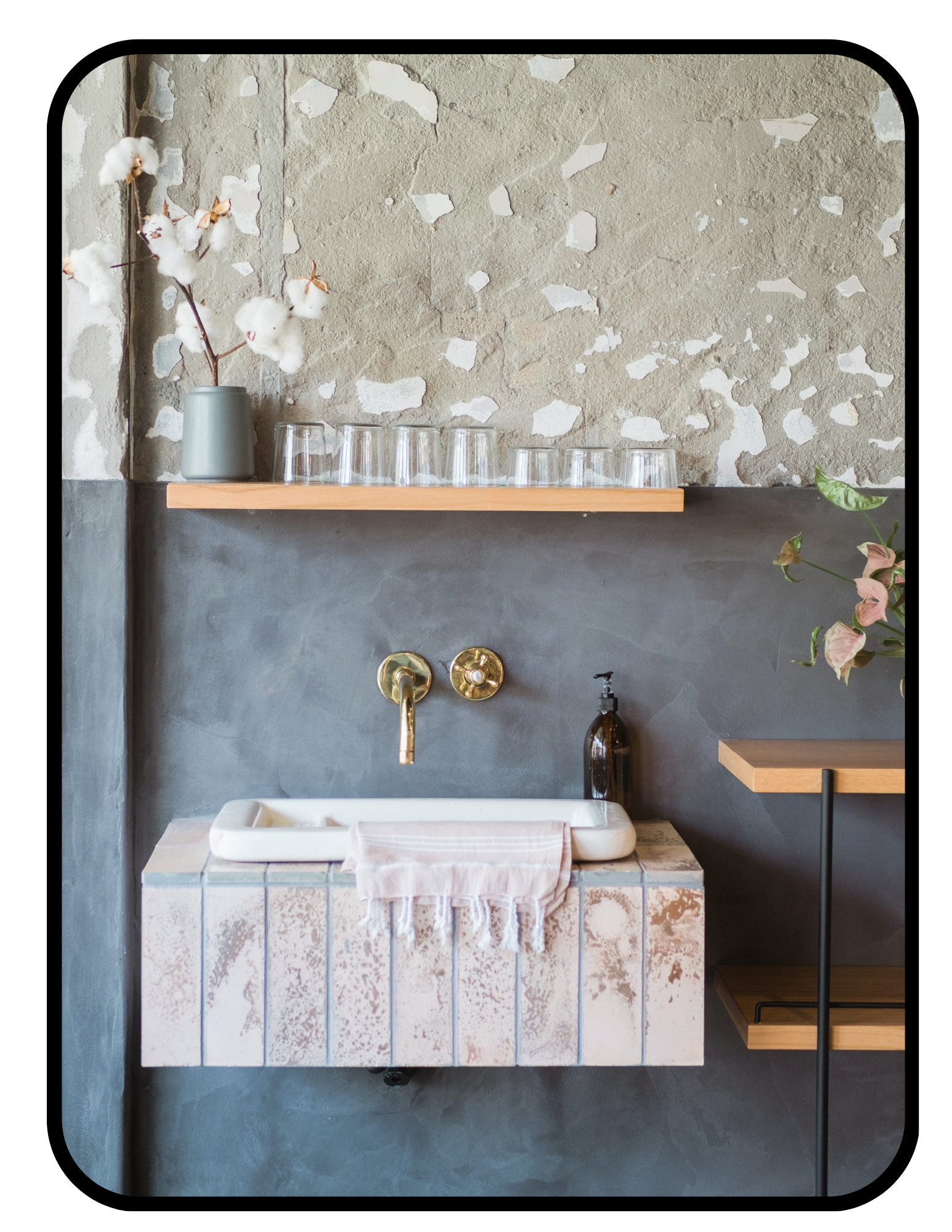 Non toxic bathroom with a stone sink, gold faucet, brown liquid soap dispenser, pink towel, wooden shelves with a gray vase holding cotton flowers, and glass cups